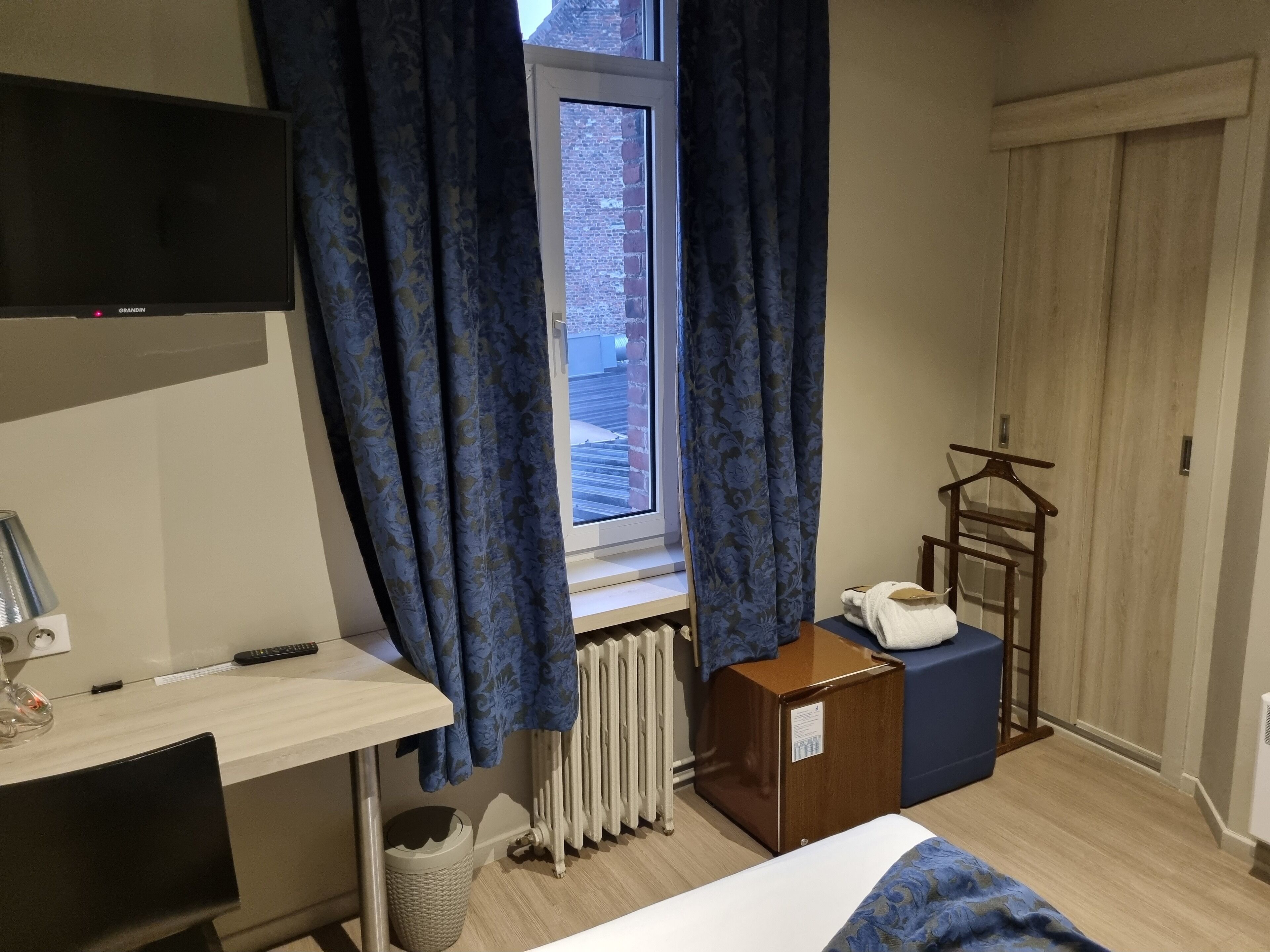 A hotel room with a bed, a desk, a chair, a radiator, a TV, and a window with curtains.
