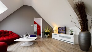A modern loft with a sloped ceiling, a dining area with a white table and red seating, a television on a white console, and a large vase with dried plants. - La Terrasse (Douai)