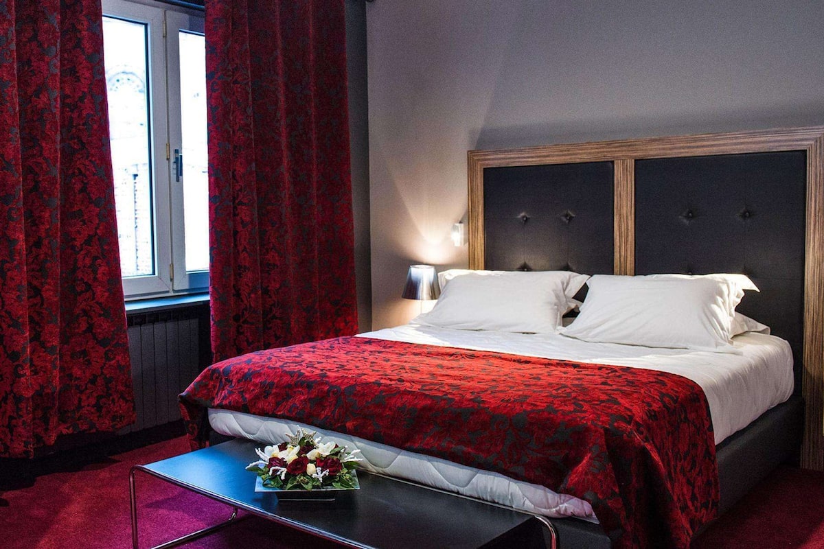 A hotel room with a bed, a bedside table with a flower arrangement, and red curtains.
