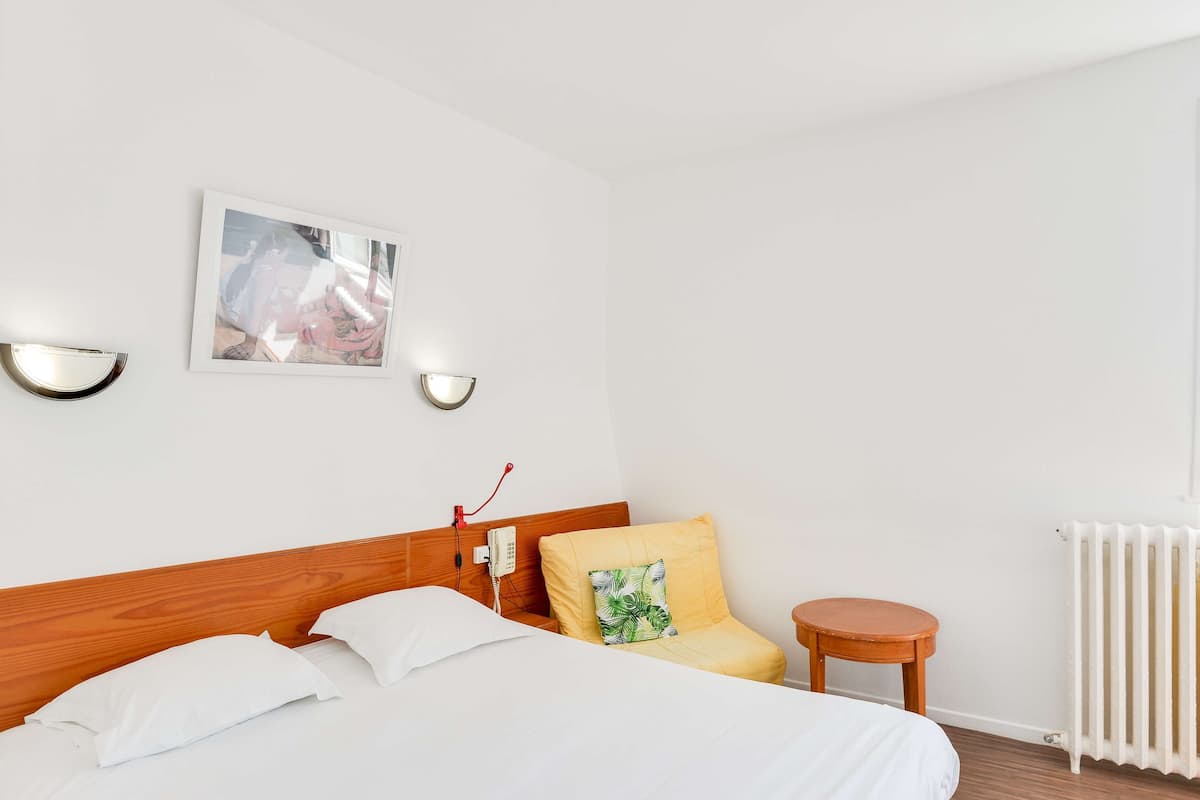 Habitación de hotel con cabecera de madera, cama con ropa blanca, almohada amarilla, una pequeña mesa redonda de madera, una lámpara de pared y un cuadro en la pared.