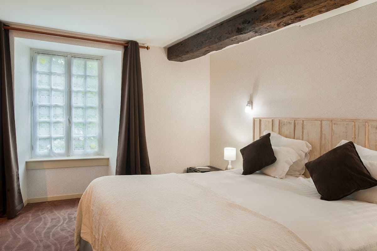 A hotel room with a large bed, a nightstand with a lamp, a window with curtains, and a wooden ceiling beam.