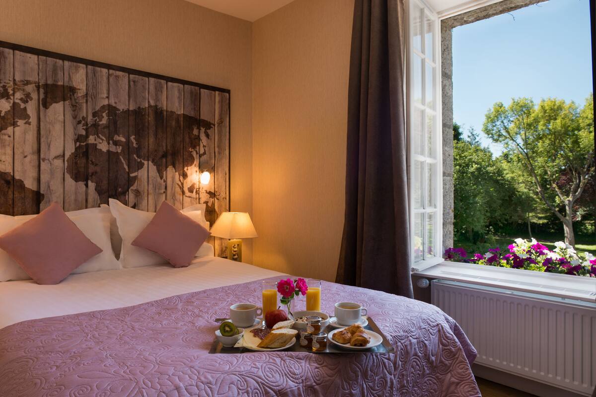 A neatly made bed with a tray of breakfast items, a world map on the wall, and a window with a view of trees and flowers.