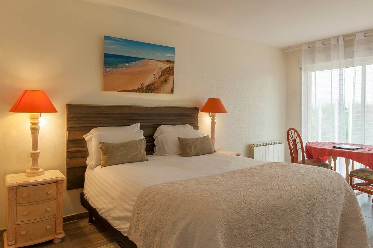 A bedroom with a bed, two bedside lamps, a painting of a beach, a table with chairs, and a window with curtains.