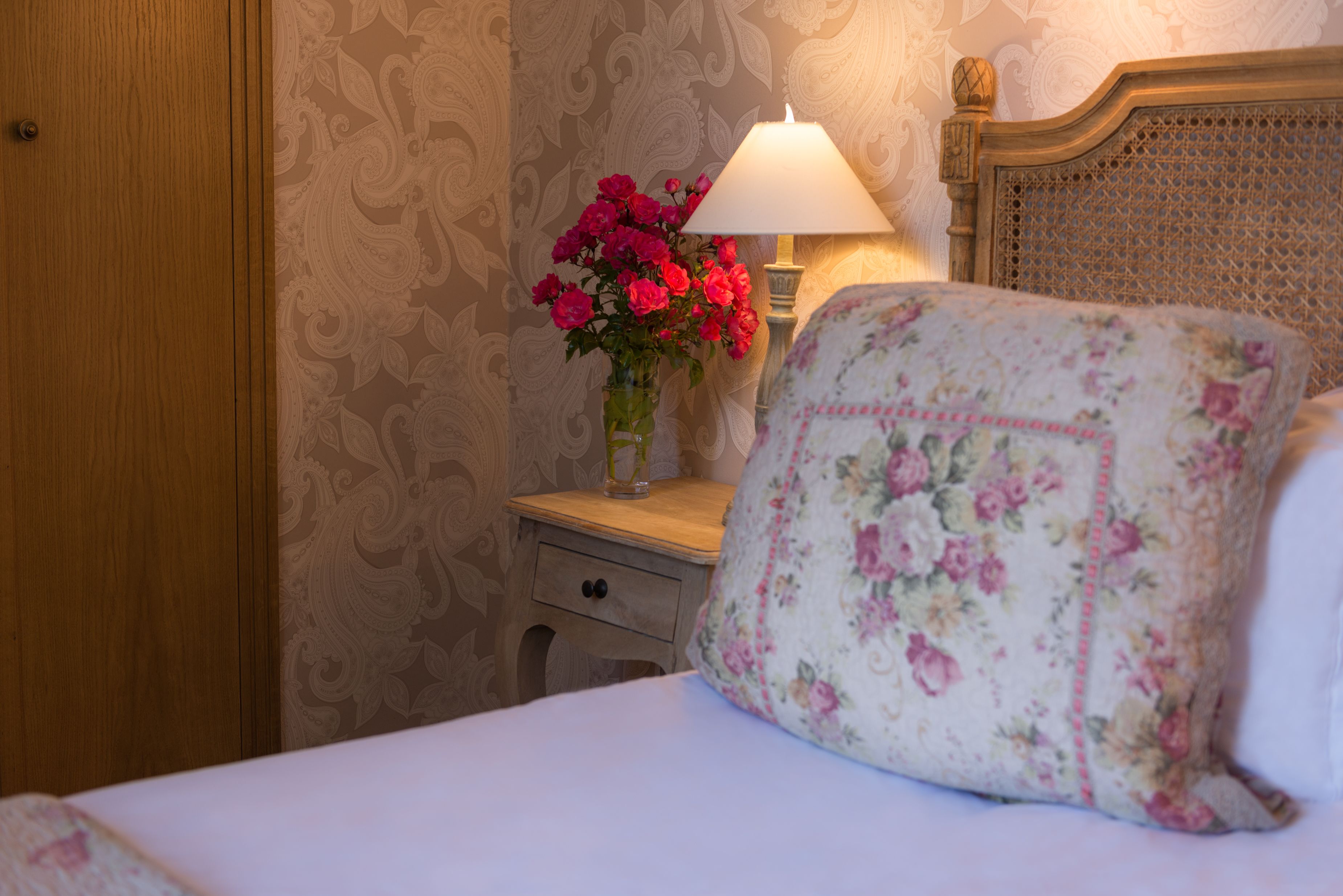 A neatly made bed with a floral pillow, a bedside table with a lamp and a vase of flowers, and a wooden wardrobe.