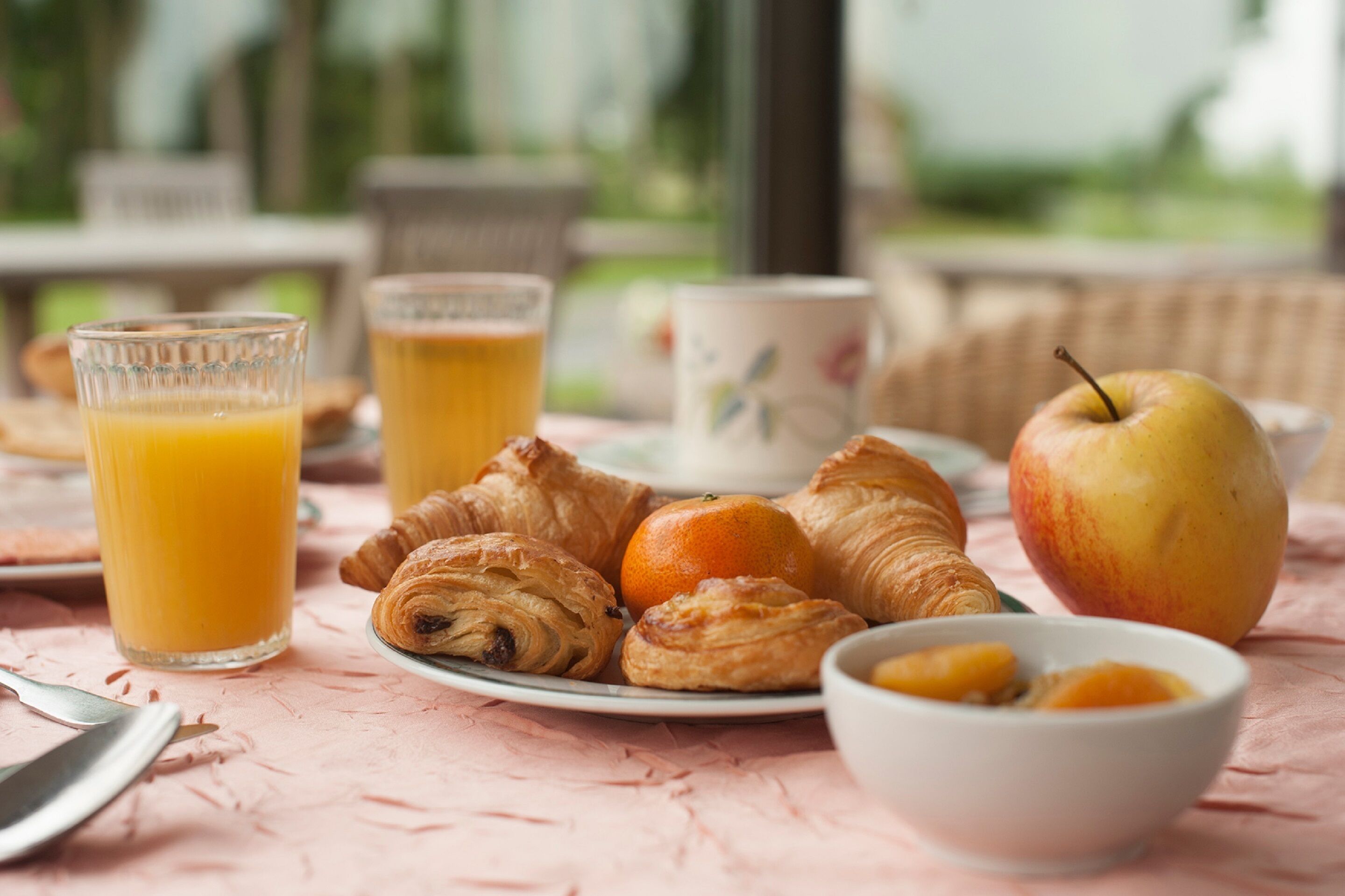 Un desayuno con croissants, una manzana, zumo de naranja y un bol de fruta sobre una mesa.