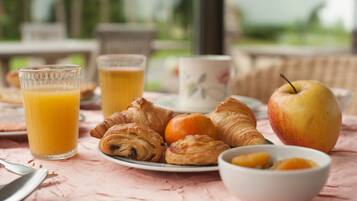 Un desayuno con croissants, una manzana, zumo de naranja y un bol de fruta sobre una mesa.