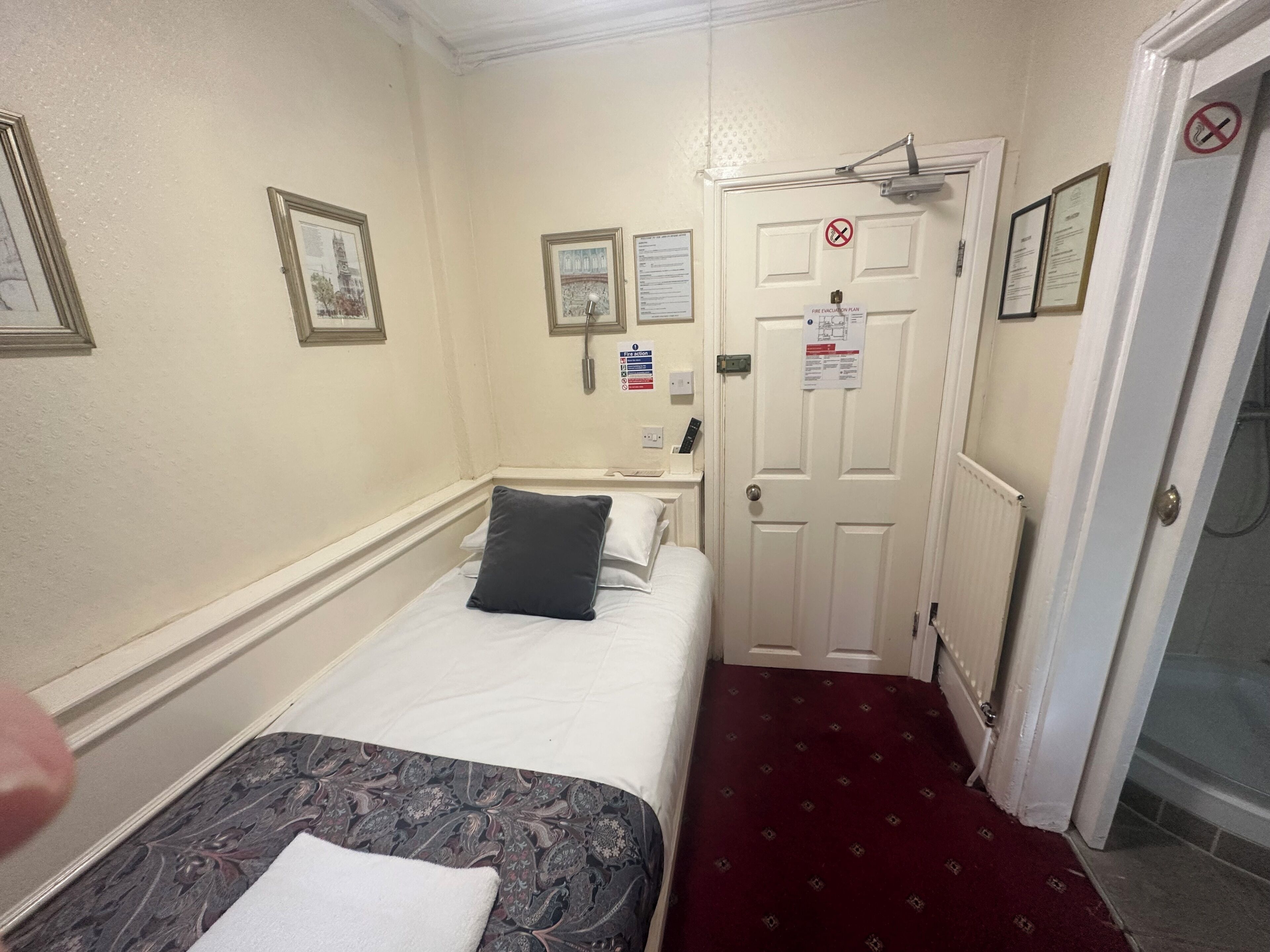 A hotel room with a bed, a bedside table, a nightstand, a door with a fire evacuation plan, and a wall-mounted telephone.