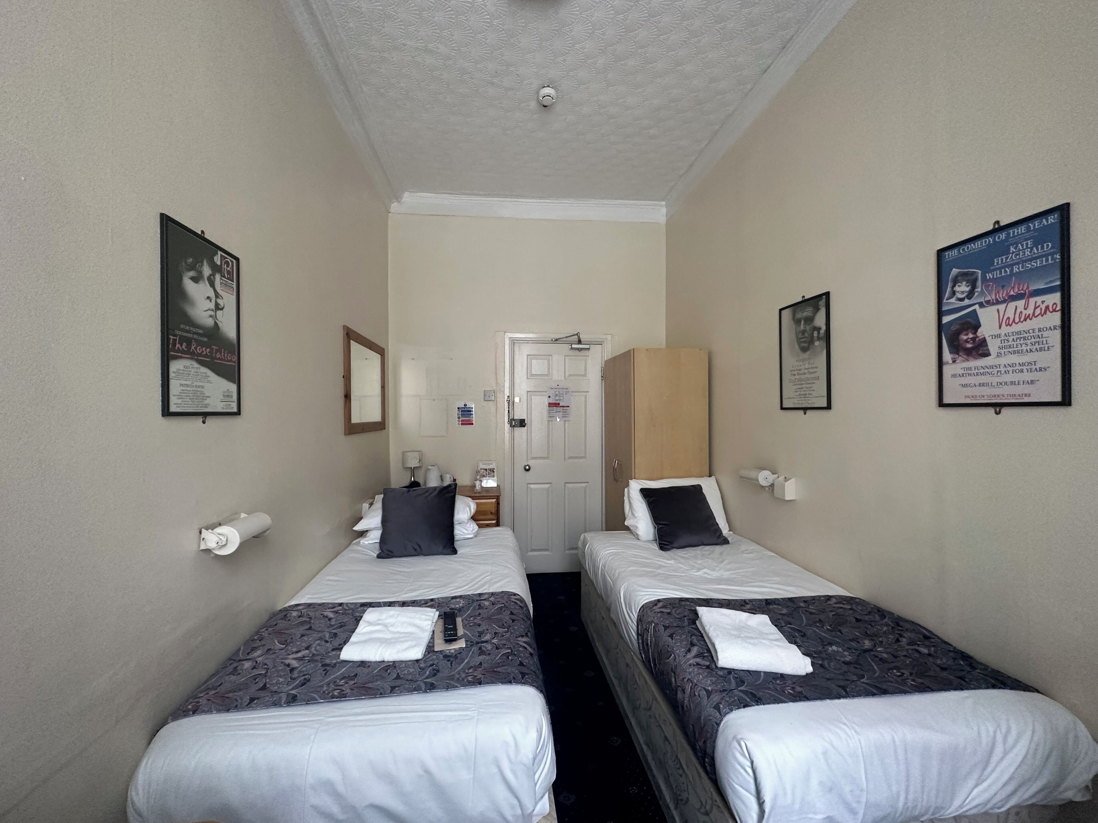 A hotel room with two single beds, a door, a mirror, a poster, and a small table with a lamp.