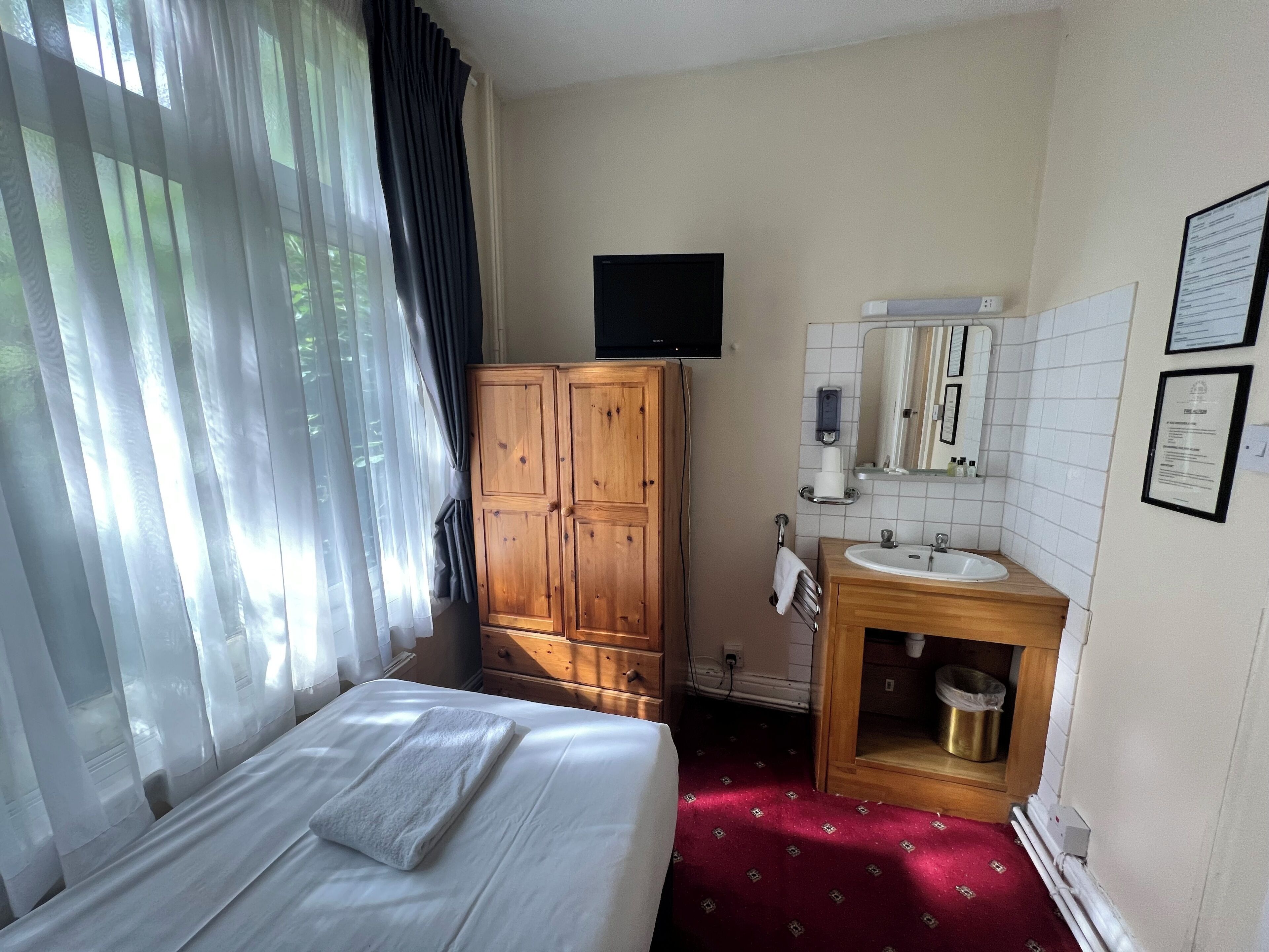 A hotel room with a bed, a wooden wardrobe, a TV mounted on the wall, a sink with a mirror, and a red carpet.