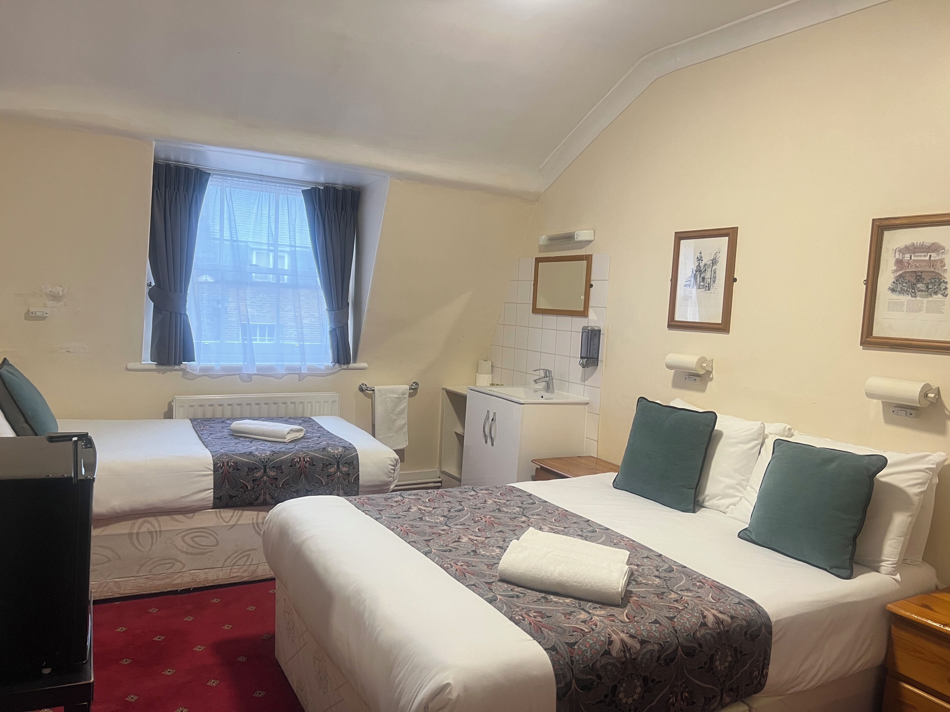 A hotel room with two beds, a window with curtains, a sink, a mirror, and framed pictures on the wall.