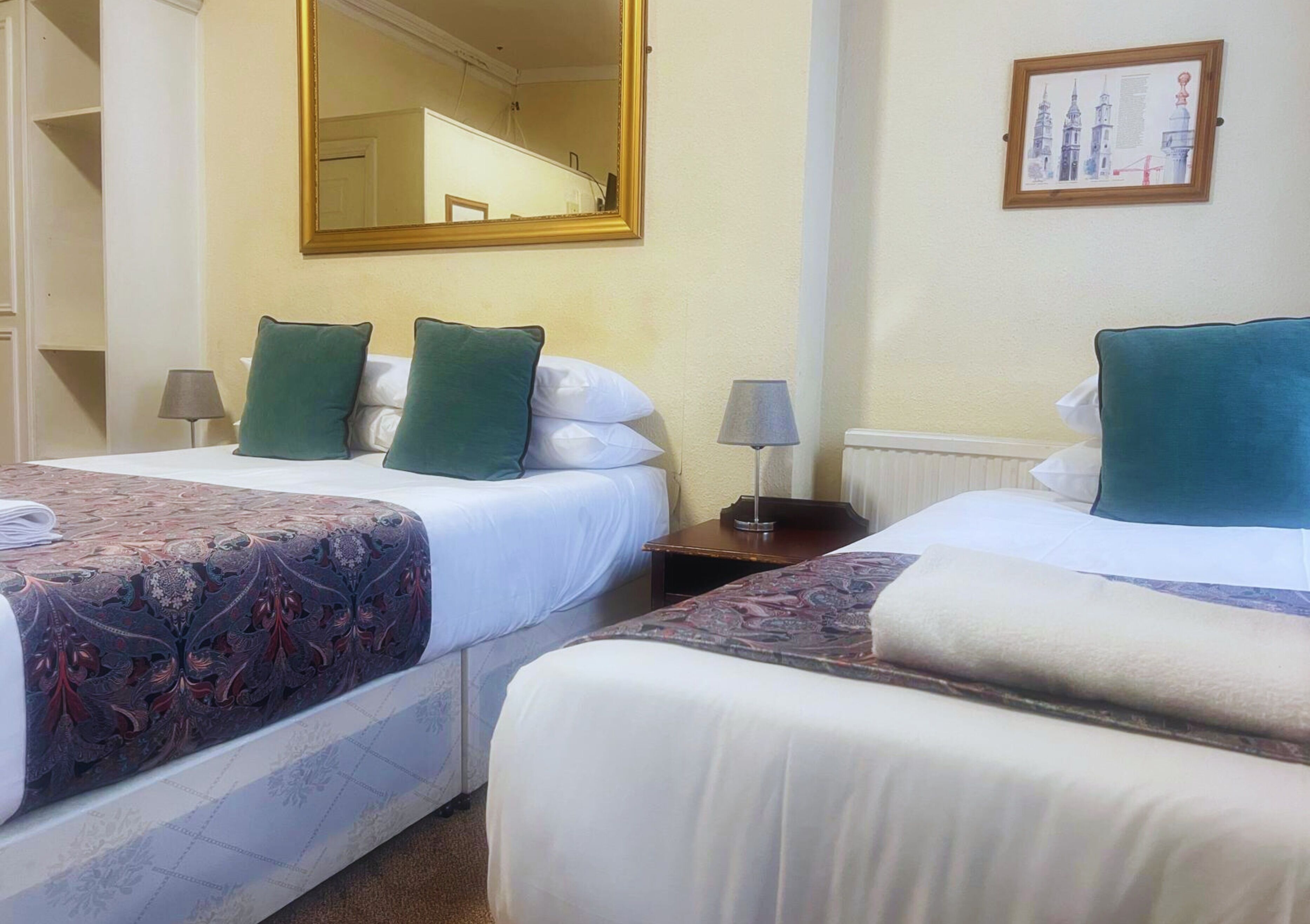 Two beds with green and white bedding, a large mirror, a framed picture on the wall, and a lamp on each bedside table.