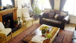 A living room with a fireplace, a leather sofa, a coffee table with fruit, and a chandelier. - Arran House Hotel (London)