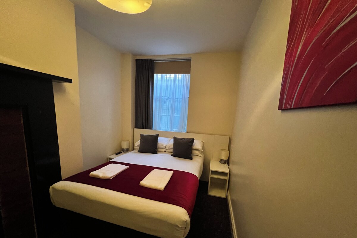 A hotel room with a bed, bedside tables, a window with curtains, and a red abstract painting on the wall.