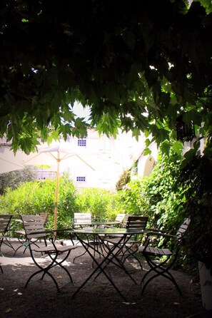 An outdoor seating area with tables and chairs, surrounded by greenery and a building in the background.