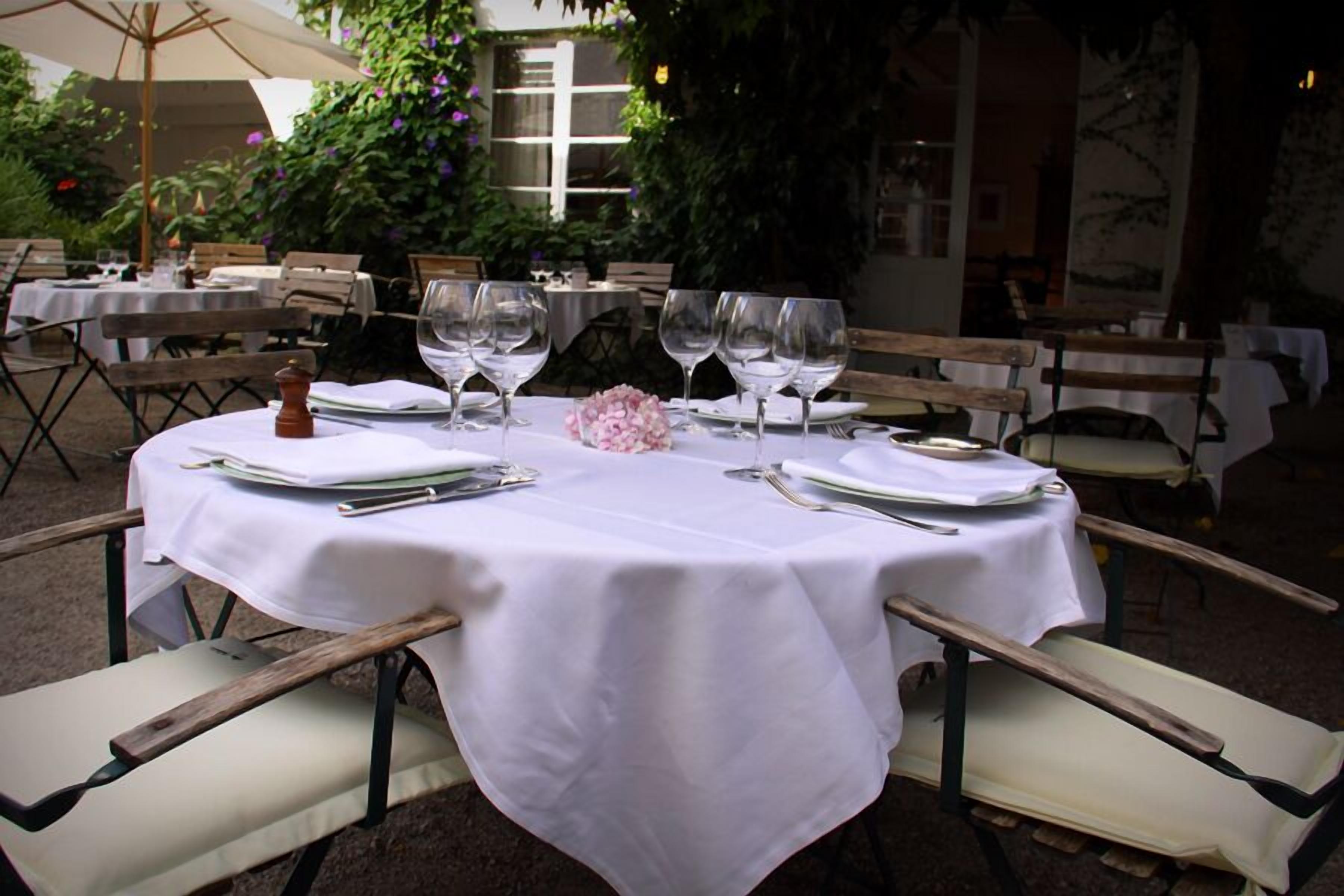An outdoor dining area with round tables set for two, each with white tablecloths, wine glasses, and cutlery.