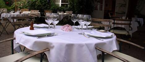 An outdoor dining area with round tables set for two, each with white tablecloths, wine glasses, and cutlery.