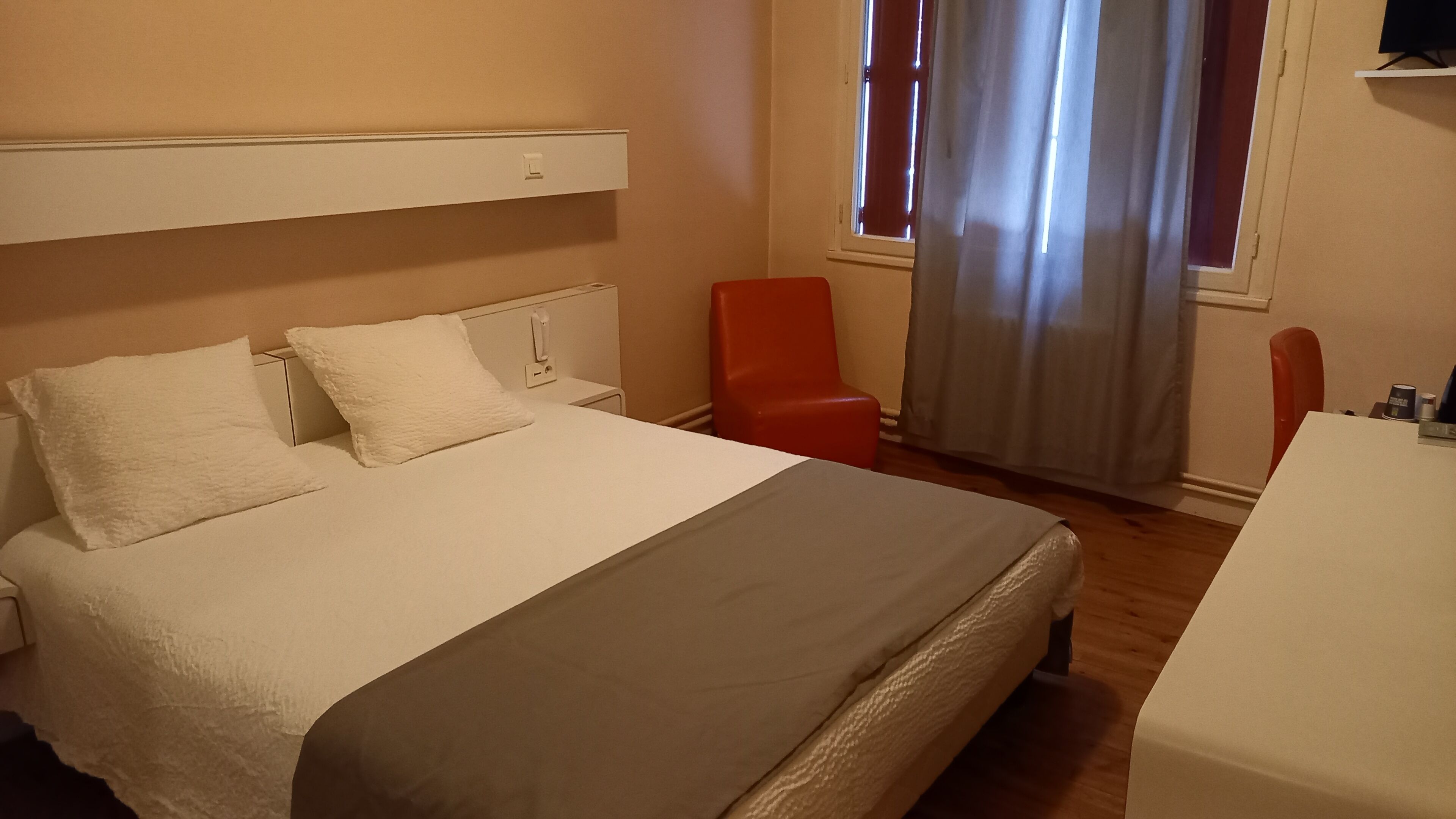 A hotel room with a bed, a red chair, a desk, and a window with curtains.