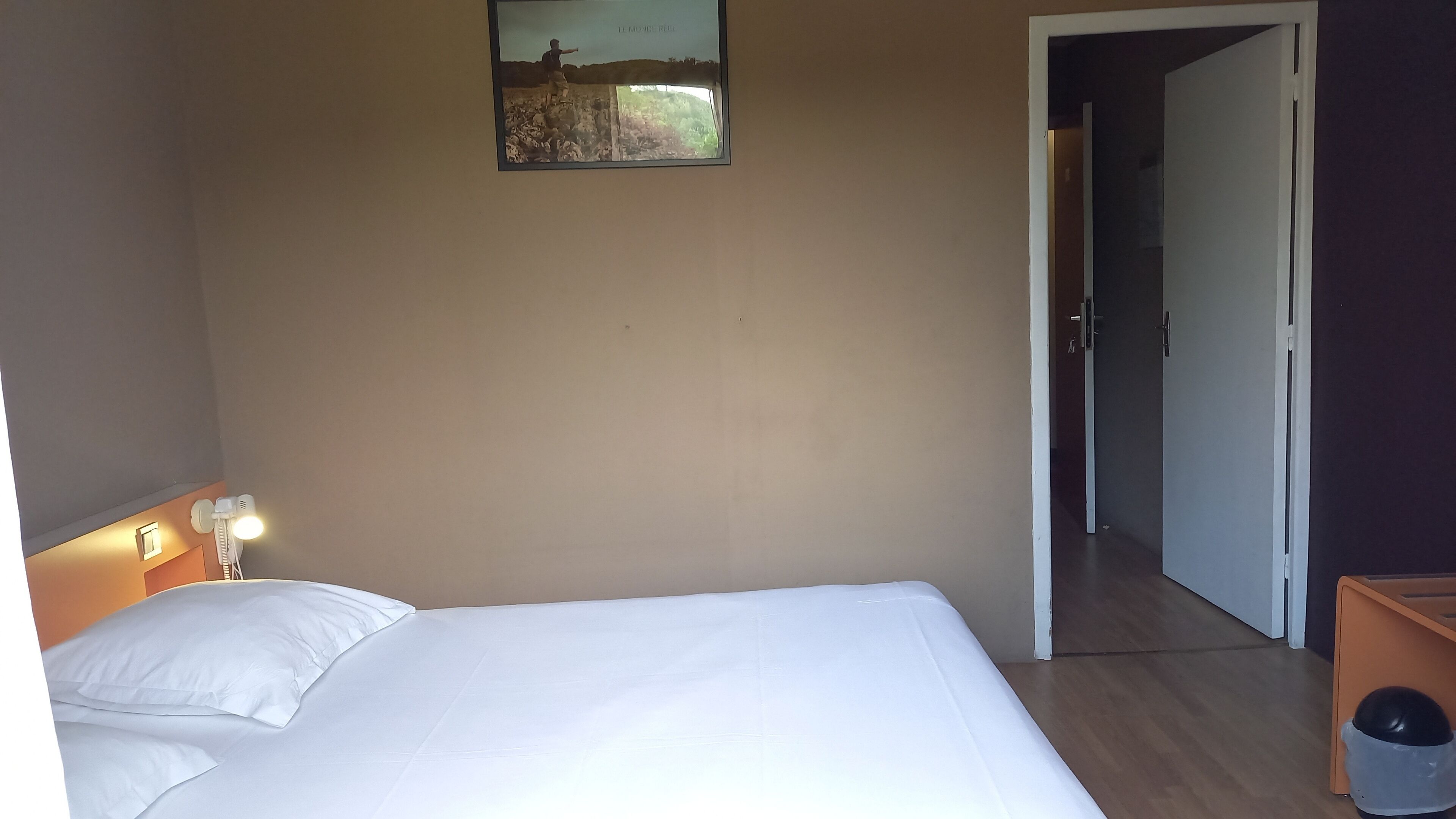 Une chambre d’hôtel avec un lit, une table de chevet avec une lampe, un tableau au mur et une porte donnant sur une autre pièce.