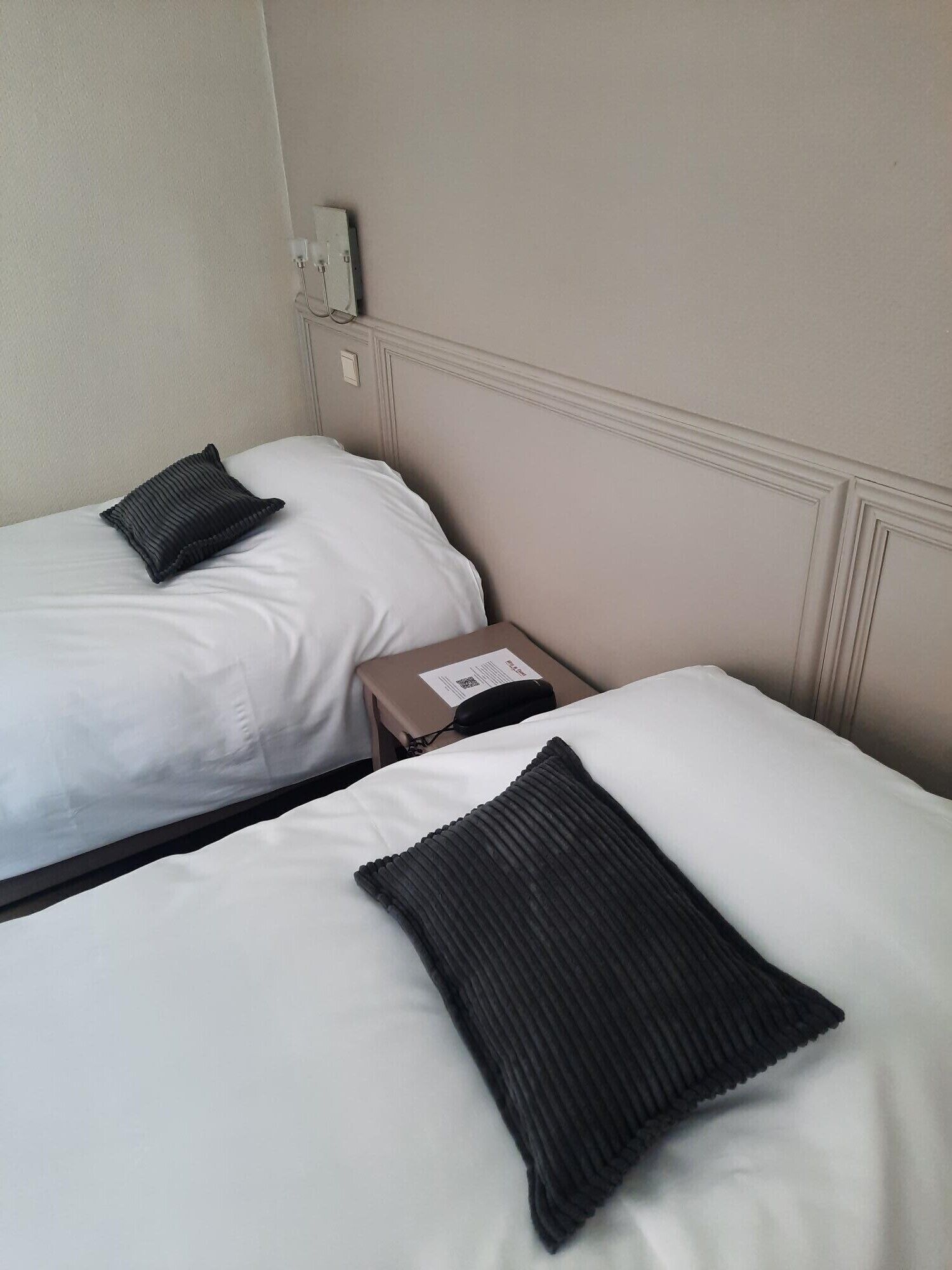 Twee eenpersoonsbedden met witte lakens en donkere kussens, een nachtkastje met een telefoon en een doos, en een wandcontactdoos.