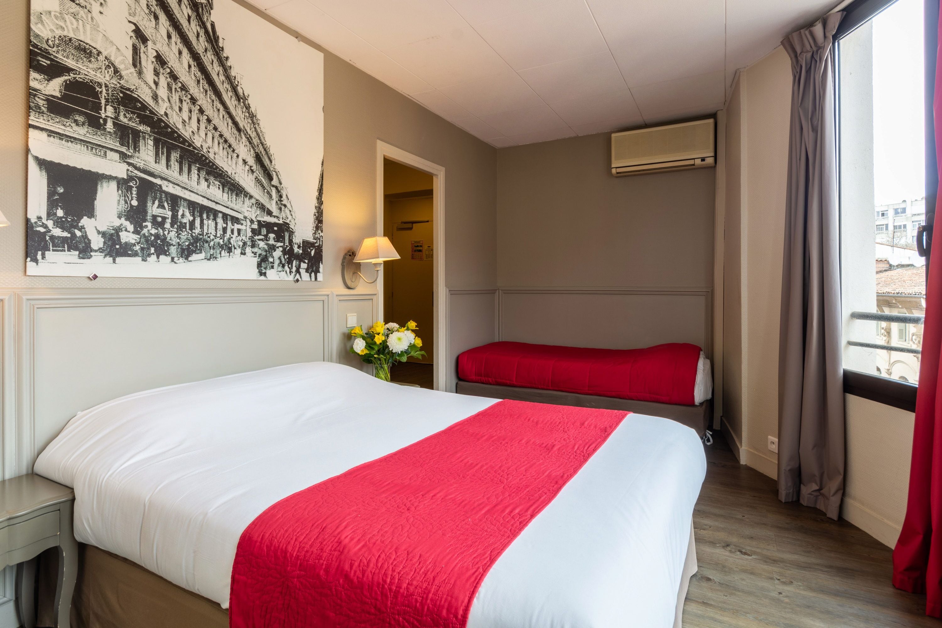 Una habitación de hotel con una cama grande, una cama más pequeña, aire acondicionado montado en la pared, una ventana con cortinas y una fotografía antigua en la pared.