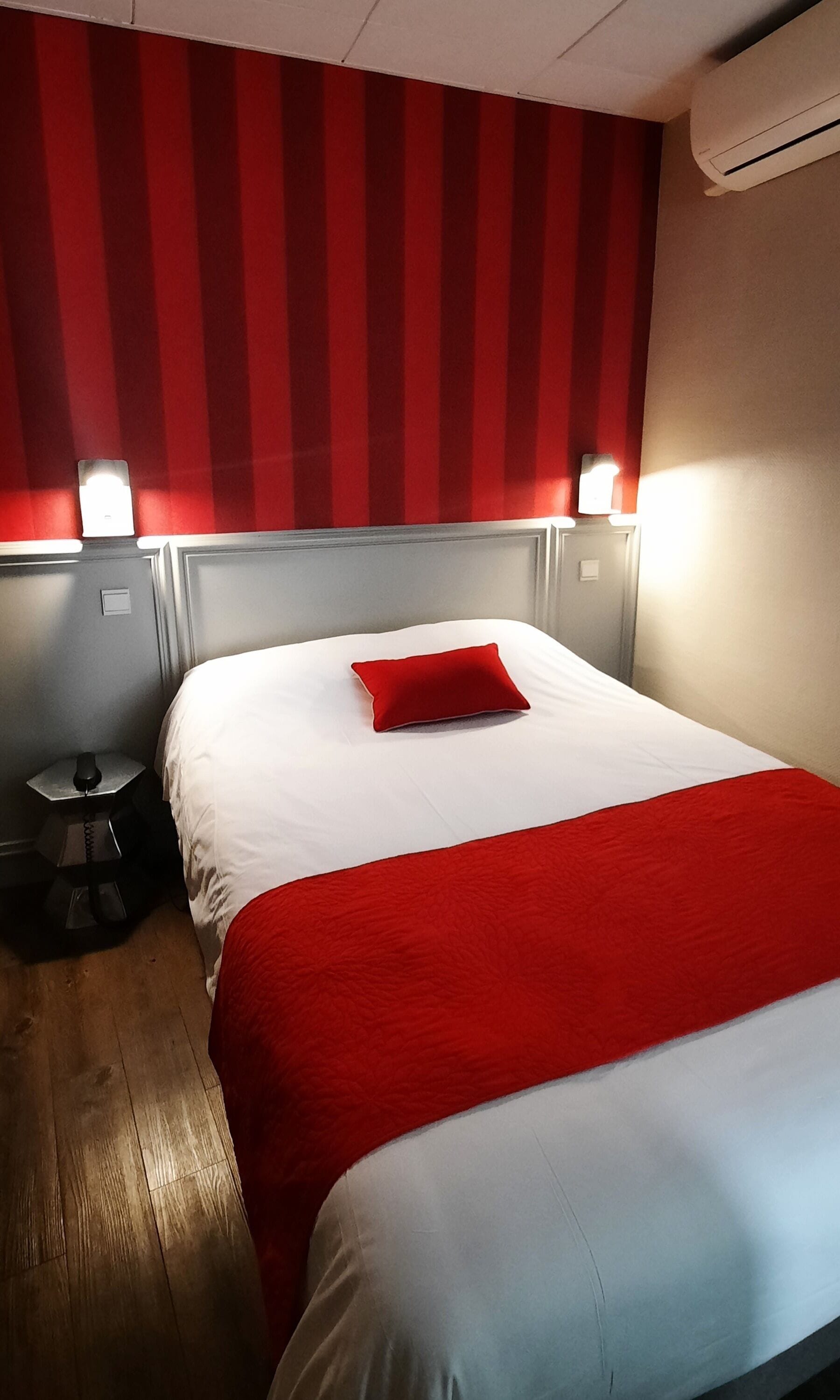 A hotel room with a bed, a red pillow, a nightstand, and a wall-mounted air conditioner.