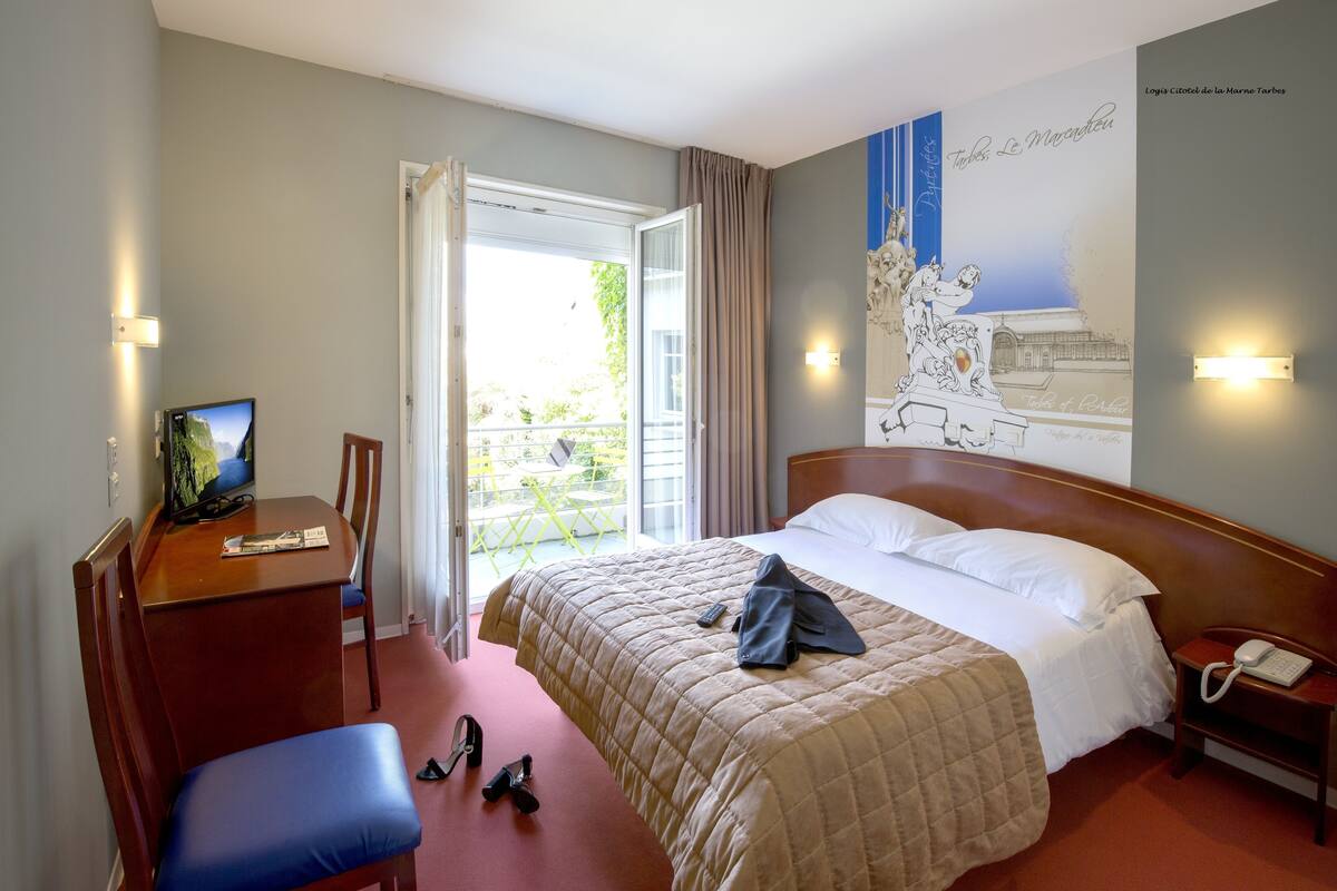 A hotel room with a bed, a desk with a computer, a chair, and a phone on a nightstand.