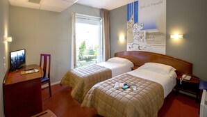 A hotel room with two beds, a desk with a TV, and a window with a view of greenery.