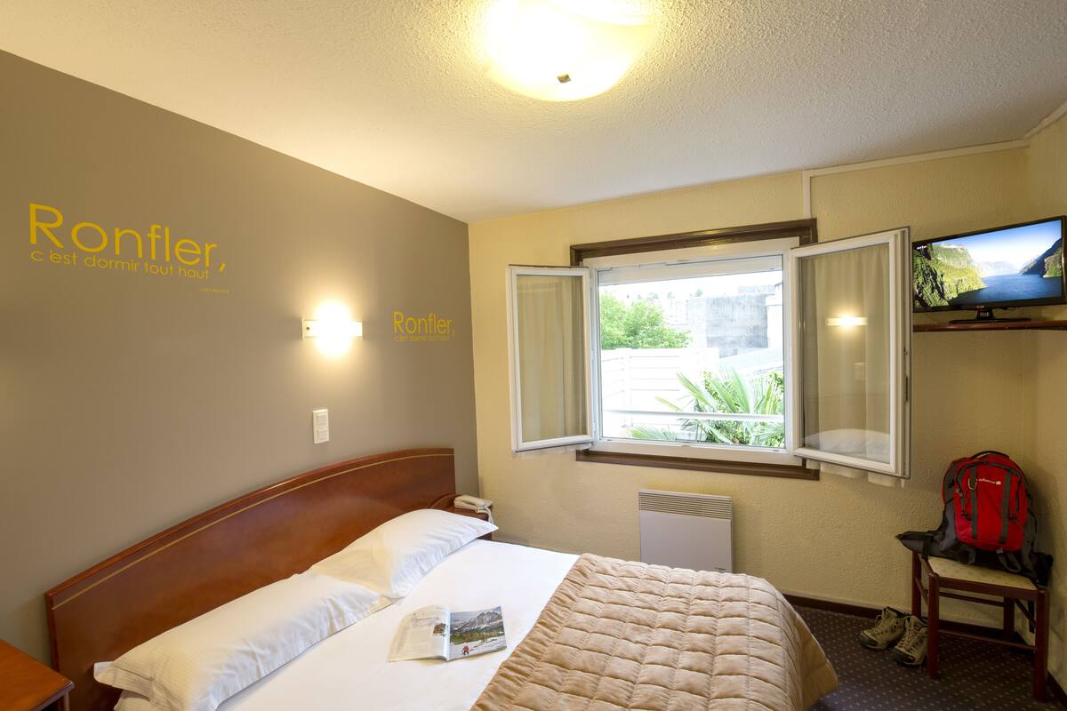 A hotel room with a bed, a window with an open view, a television on a stand, and a red backpack on a chair.