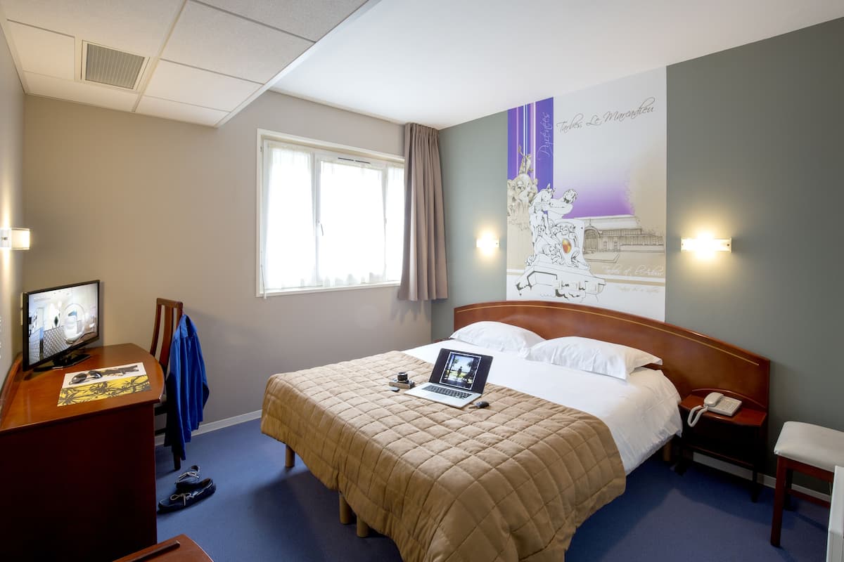 A hotel room with a bed, a desk with a computer, a chair, and a window with curtains.