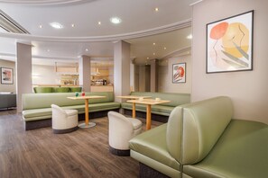 Un'area lounge moderna con sedute verdi, tavoli di legno e una cucina sullo sfondo.