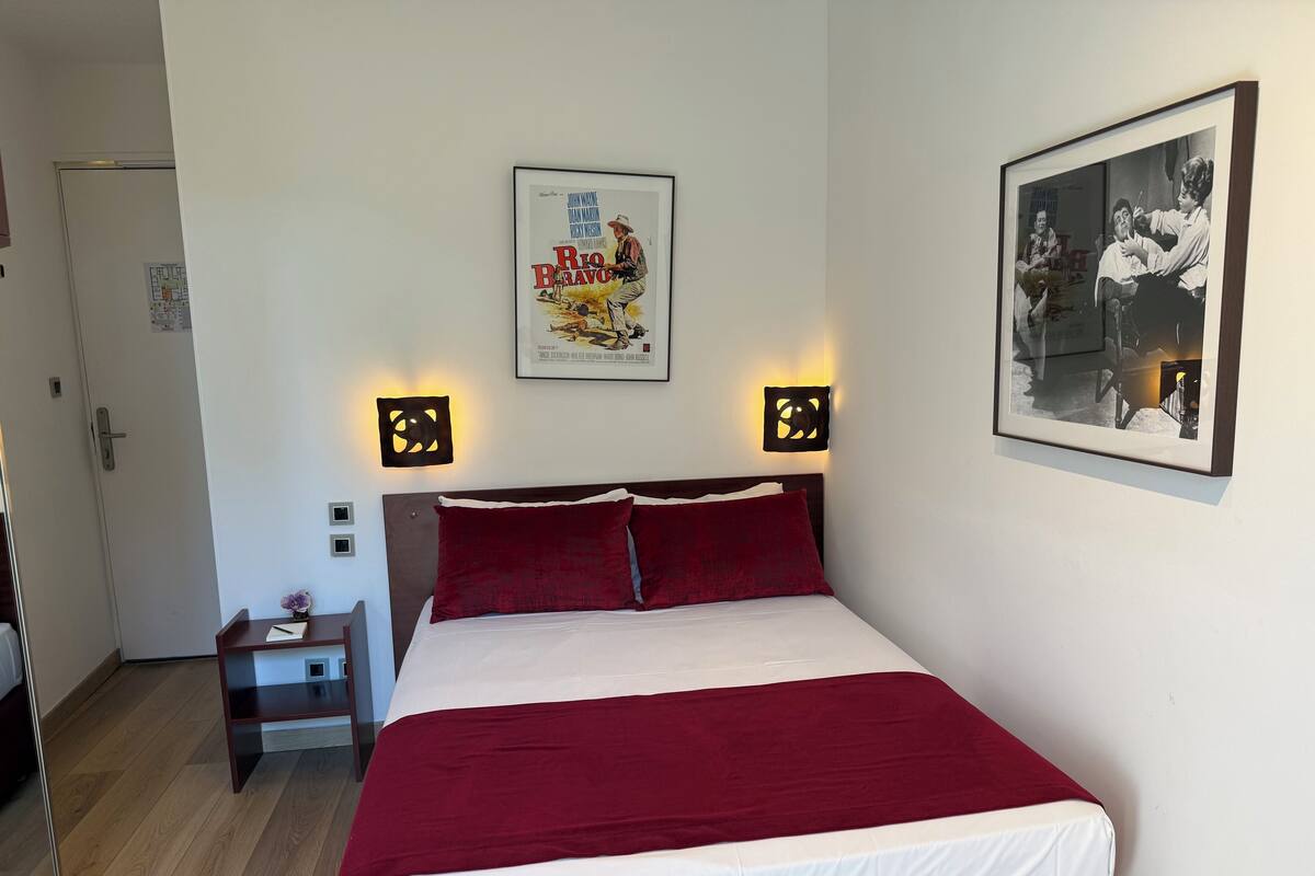 A hotel room with a bed, a nightstand, a framed movie poster, and a wall-mounted light fixture.