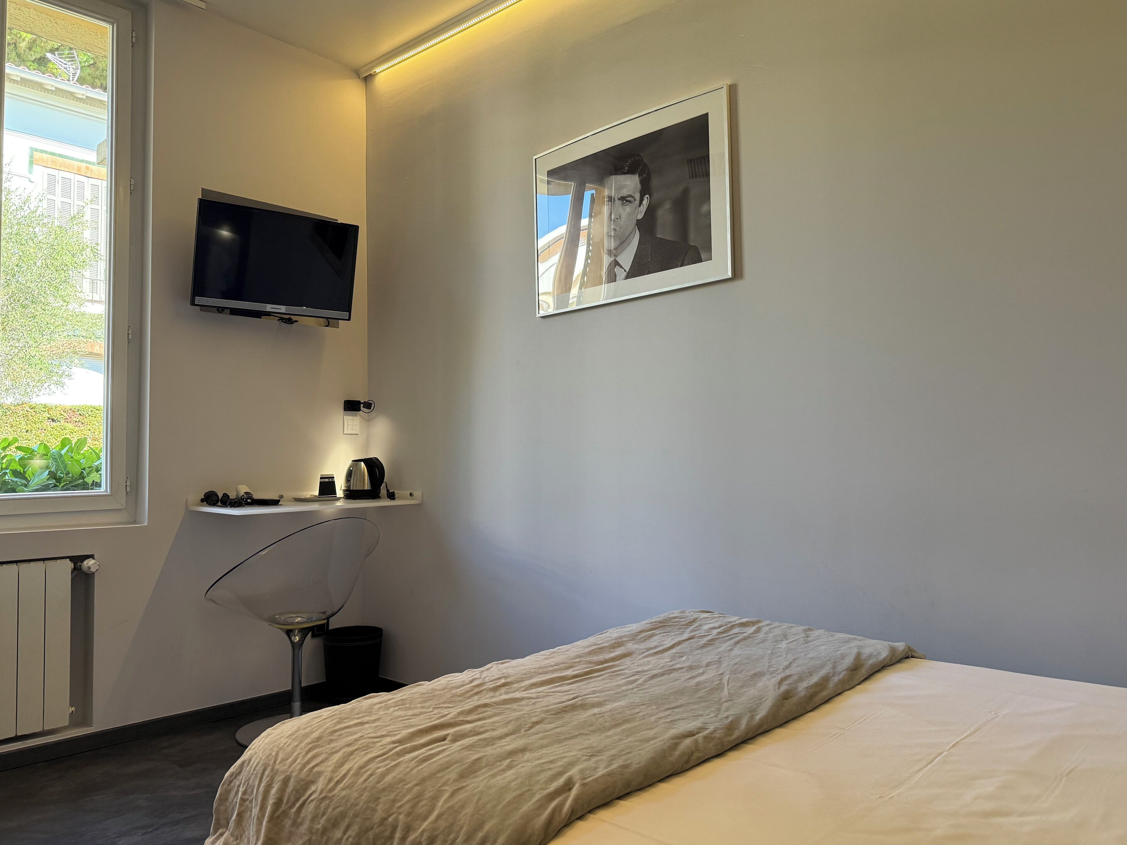 A bedroom with a bed, a TV mounted on the wall, a small desk with a kettle, and a window with a view of greenery.