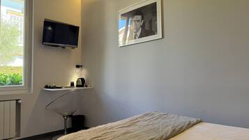 A bedroom with a bed, a TV mounted on the wall, a small desk with a kettle, and a window with a view of greenery.