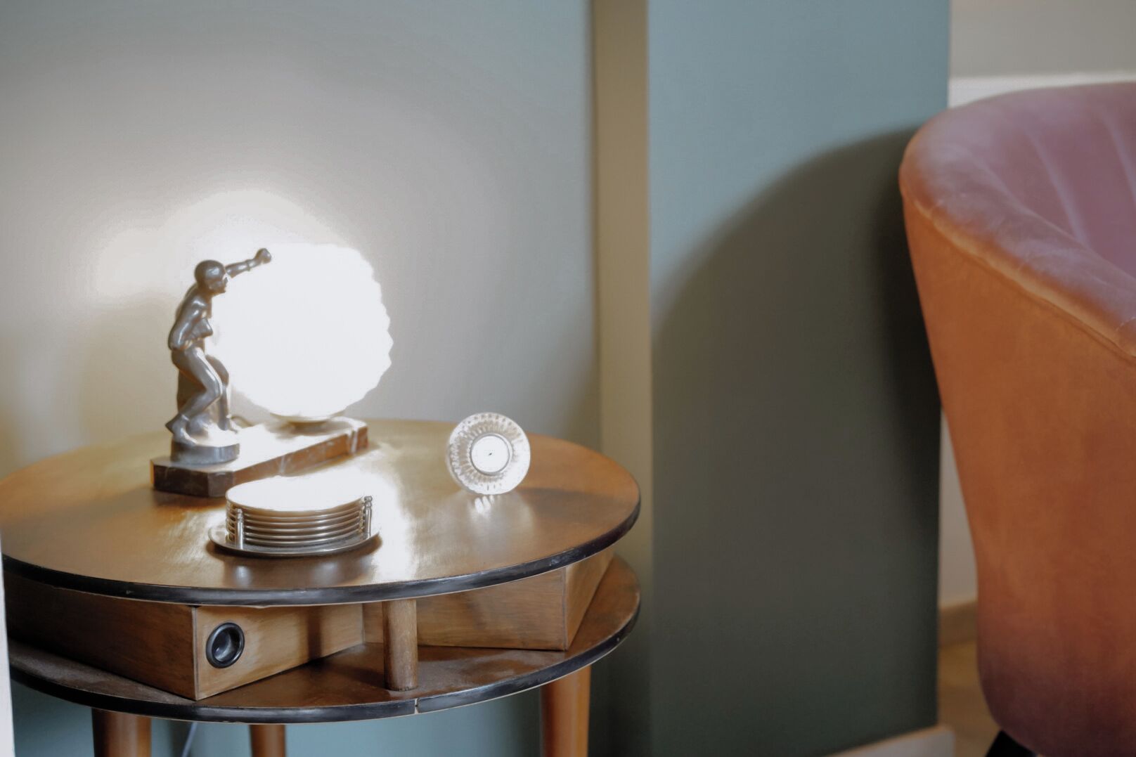 A round wooden side table with a statue, a stack of plates, and a glass object.