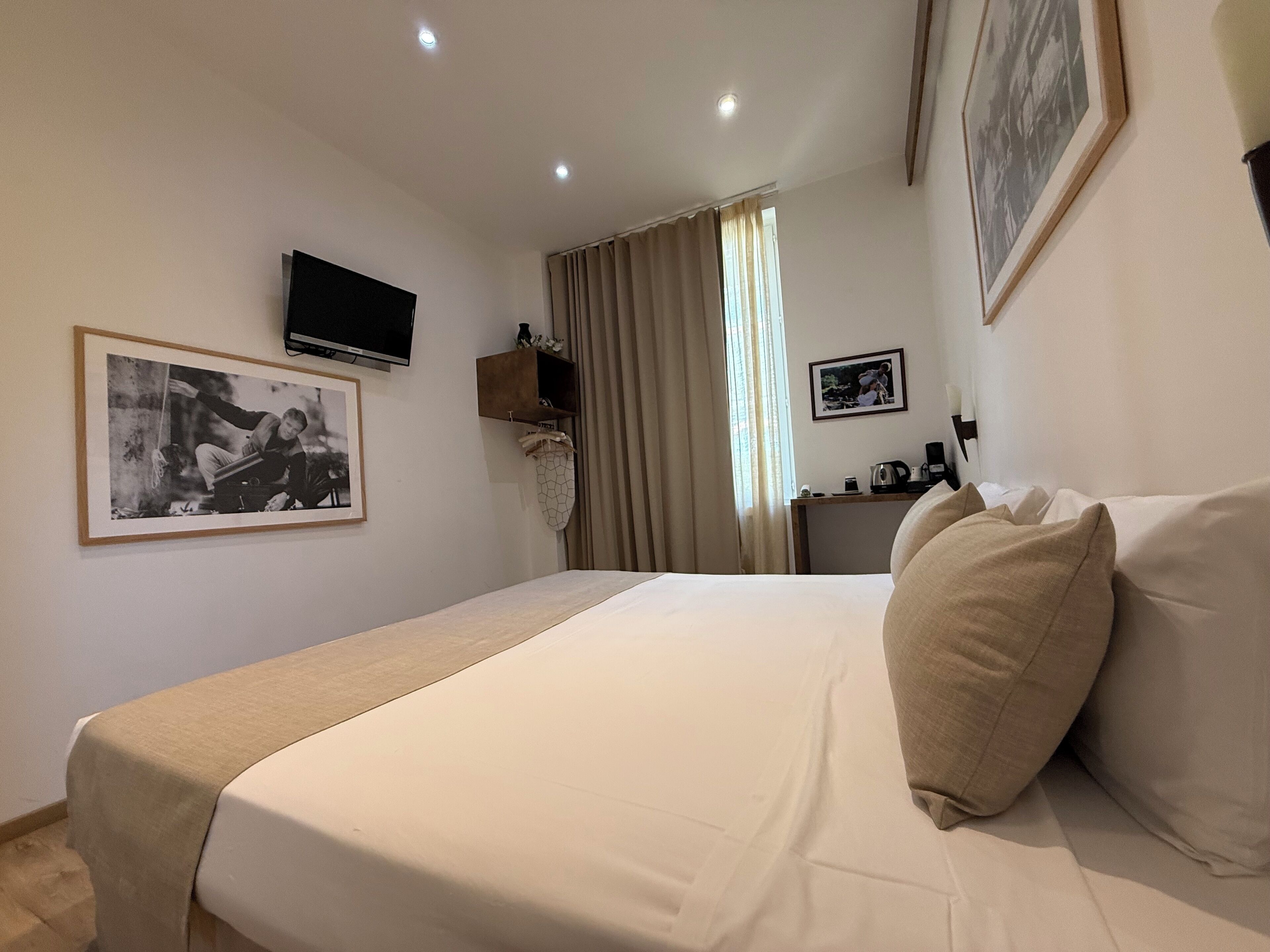 A hotel room with a bed, a TV mounted on the wall, a desk with a chair, and a window with curtains.