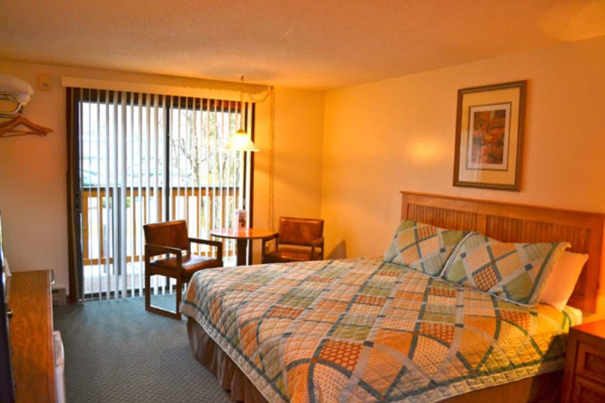 A hotel room with a bed, a chair, a small table, and a window with blinds.