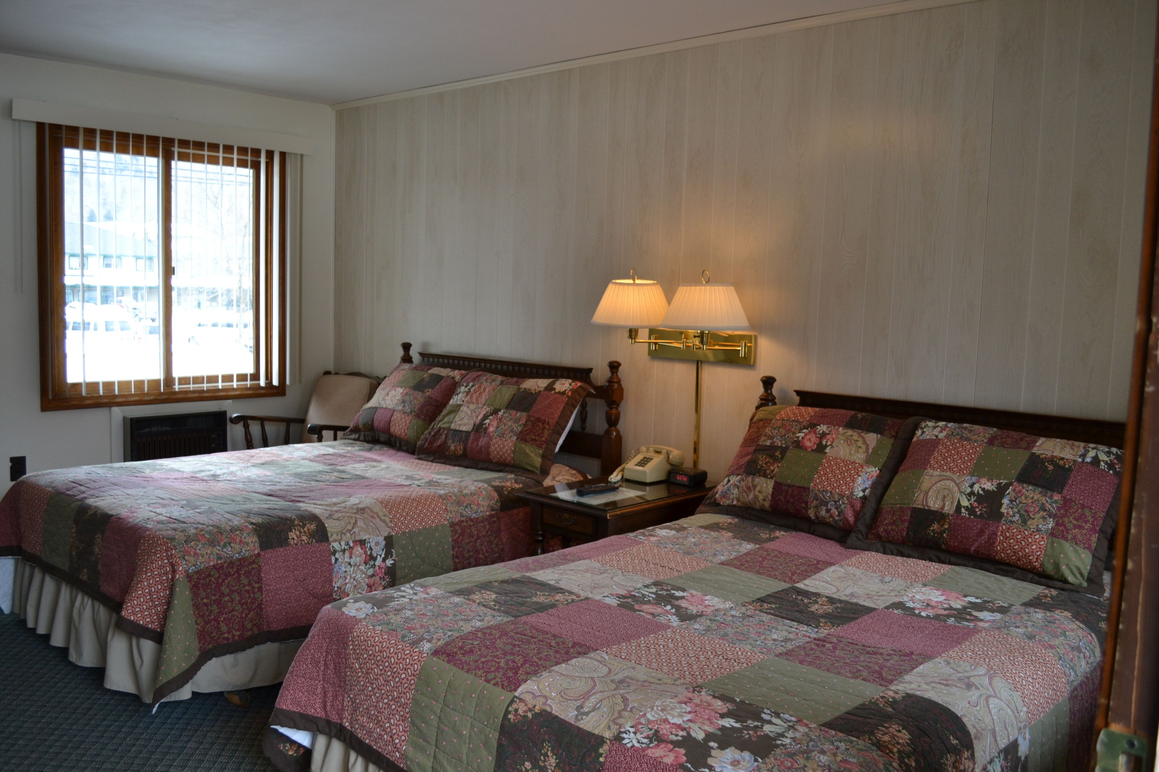 A hotel room with two beds, a nightstand with a lamp, a chair, and a window with blinds.