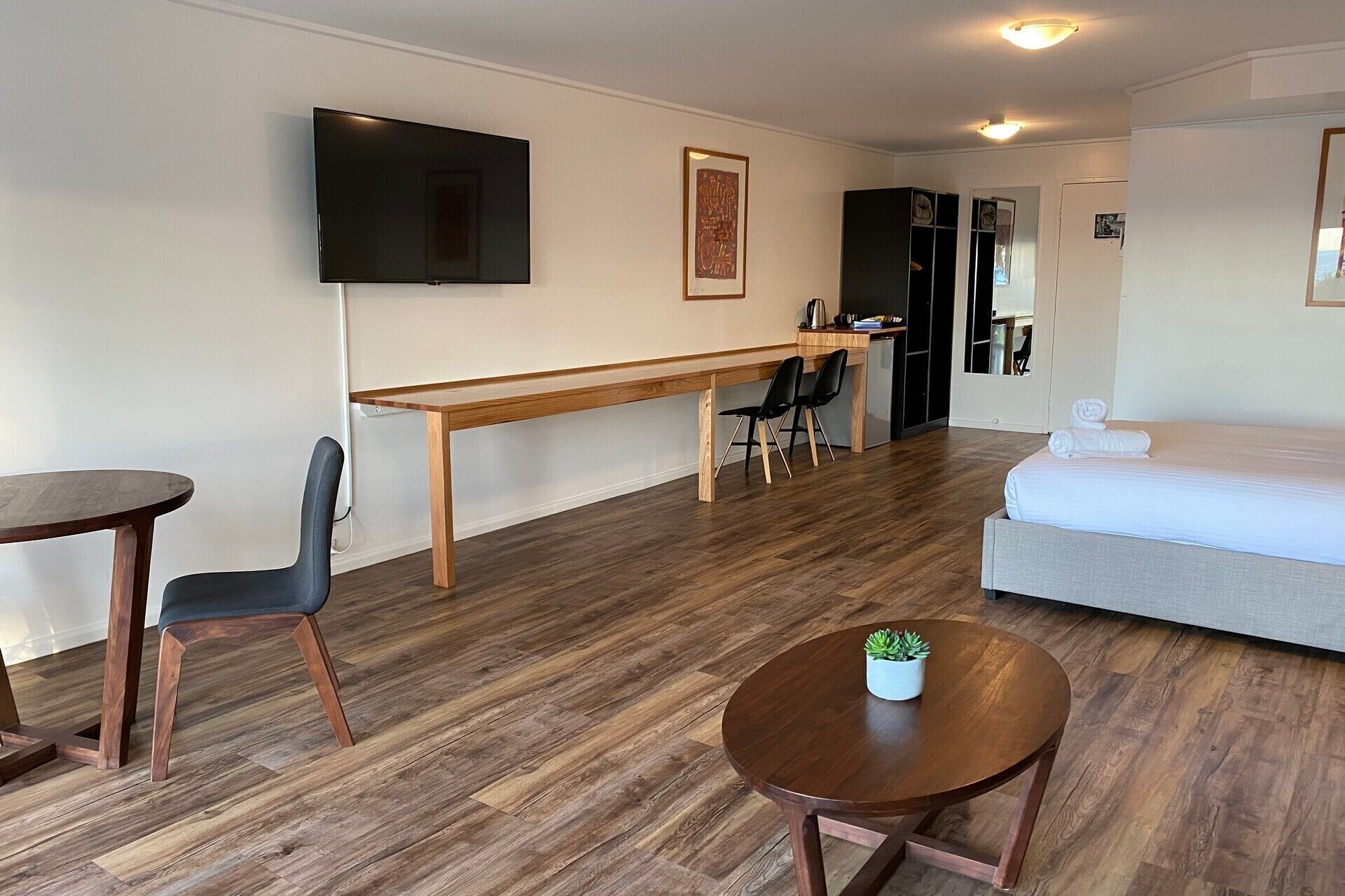 Una habitación de hotel moderna con una cama, un escritorio, dos sillas, una mesita con una planta y un televisor en la pared.
