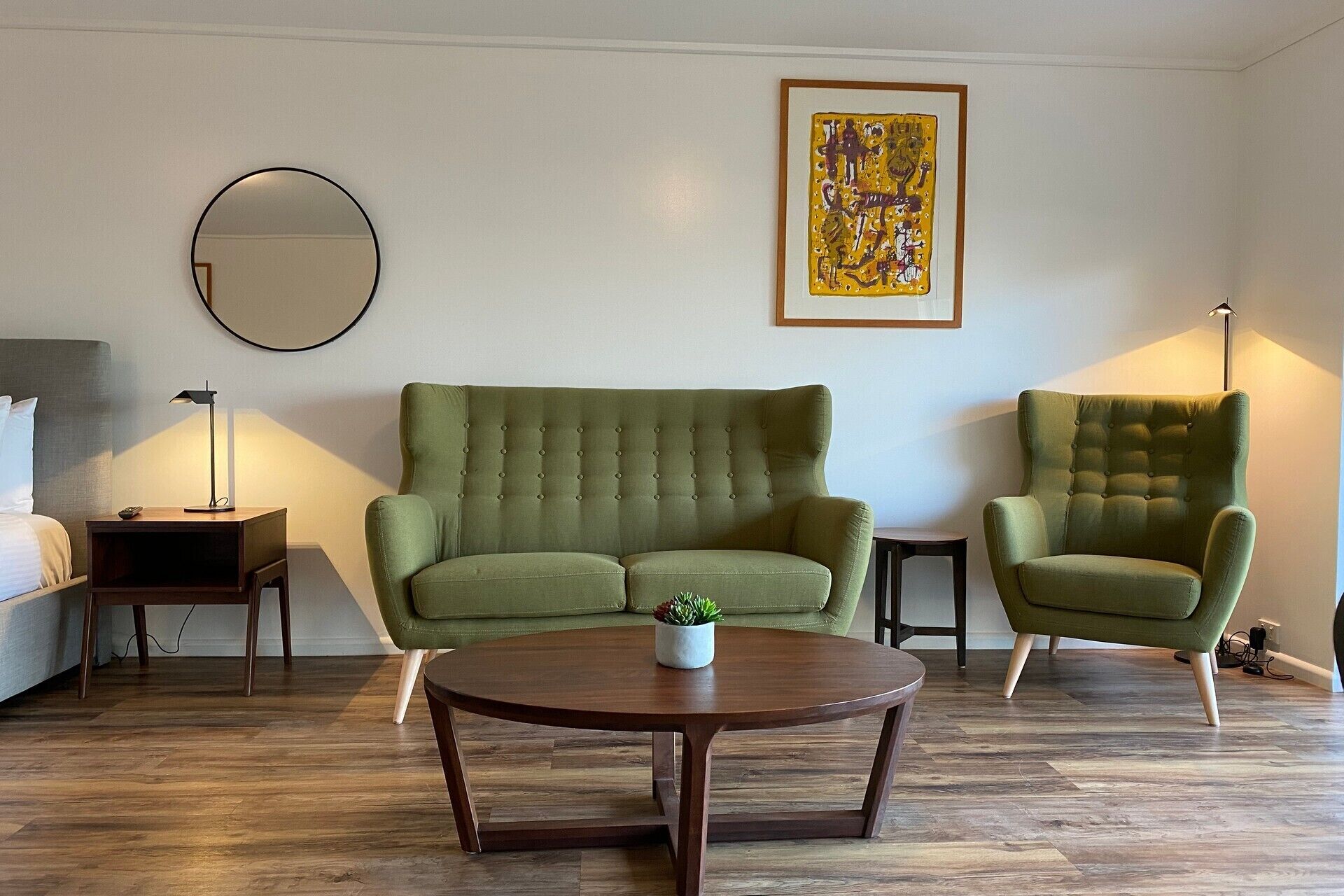 Un salón con un sofá verde, un sillón a juego, una mesa de centro redonda, una mesita auxiliar, una lámpara de pie, un espejo circular y una obra de arte enmarcada en la pared.