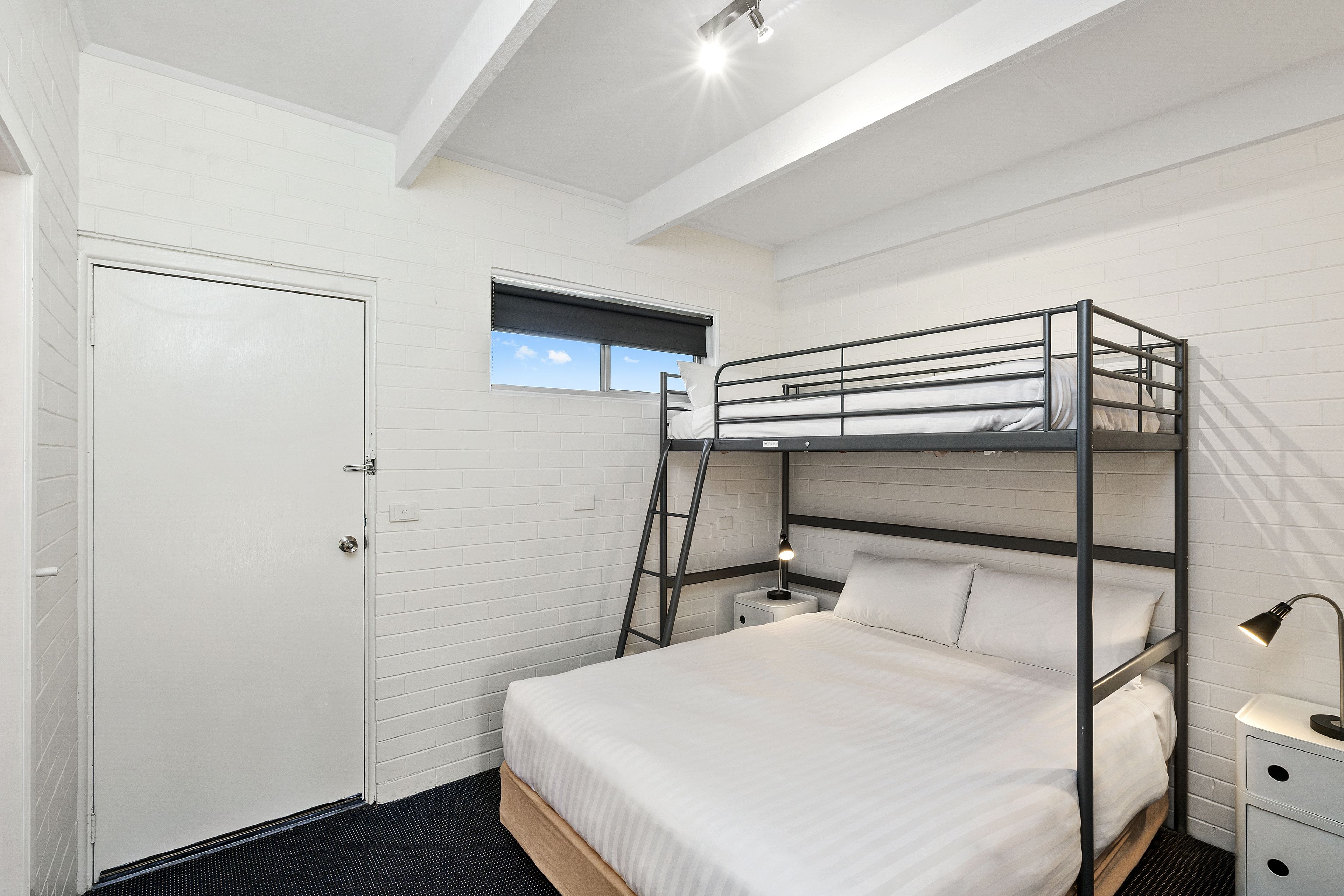 A bunk bed room with a window, a door, and a bedside table with a lamp.
