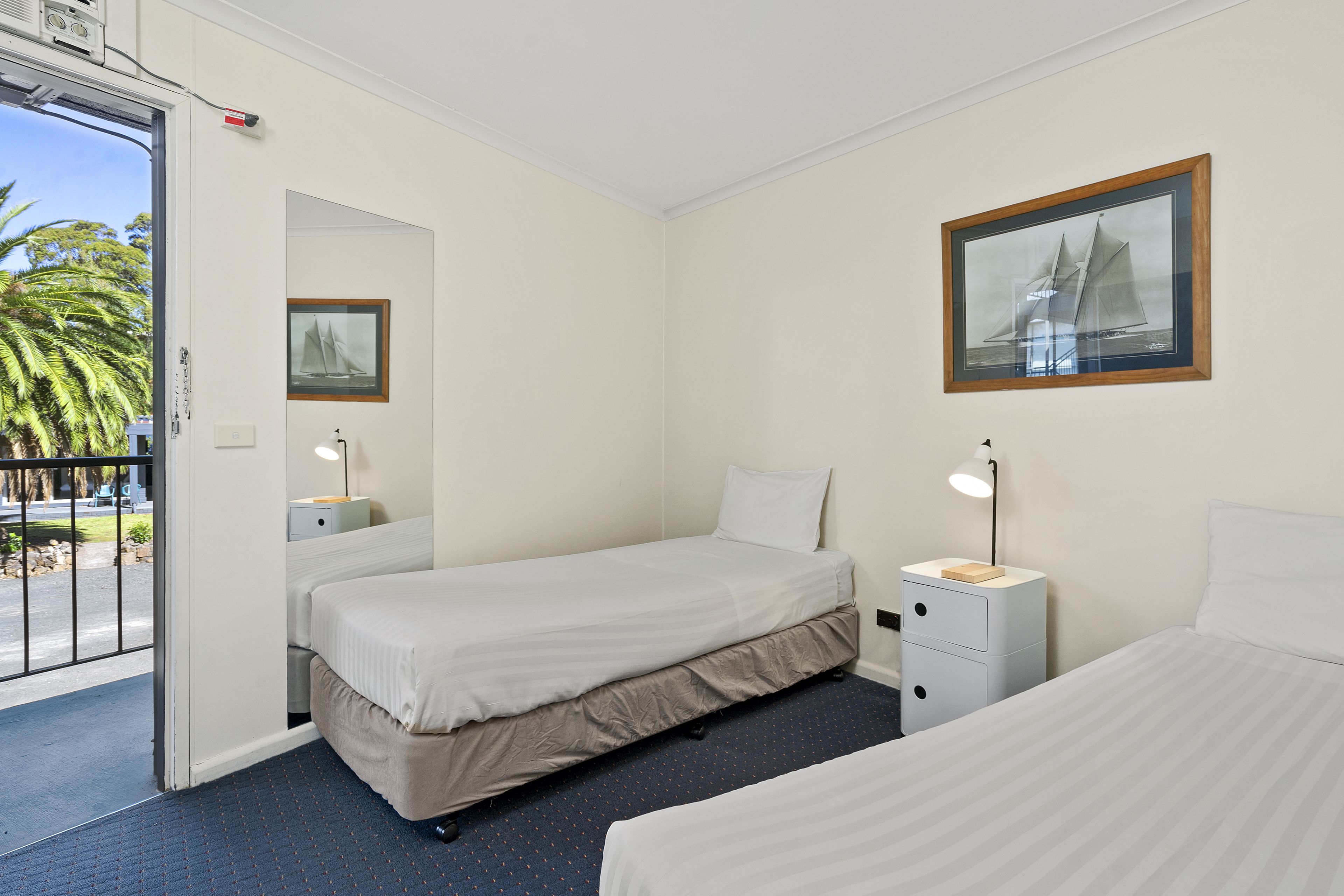 A hotel room with two beds, a small desk, a nightstand, a framed picture on the wall, and a door leading to a balcony with a view of palm trees.