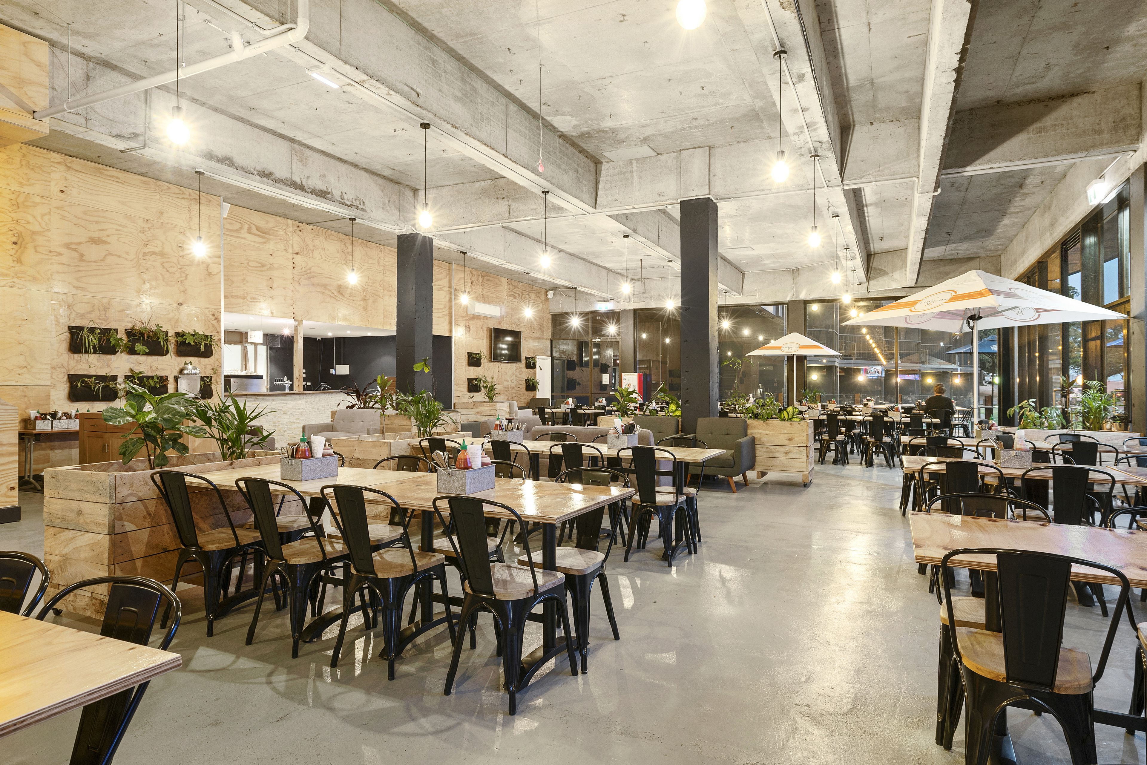 A spacious cafe with wooden tables and black chairs, large windows, and indoor plants.