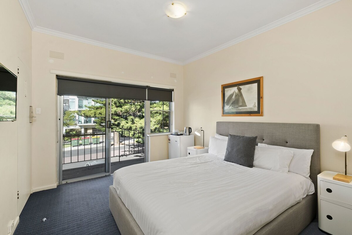 A hotel room with a bed, bedside tables, a nightstand, a framed picture, and a sliding door to the balcony.