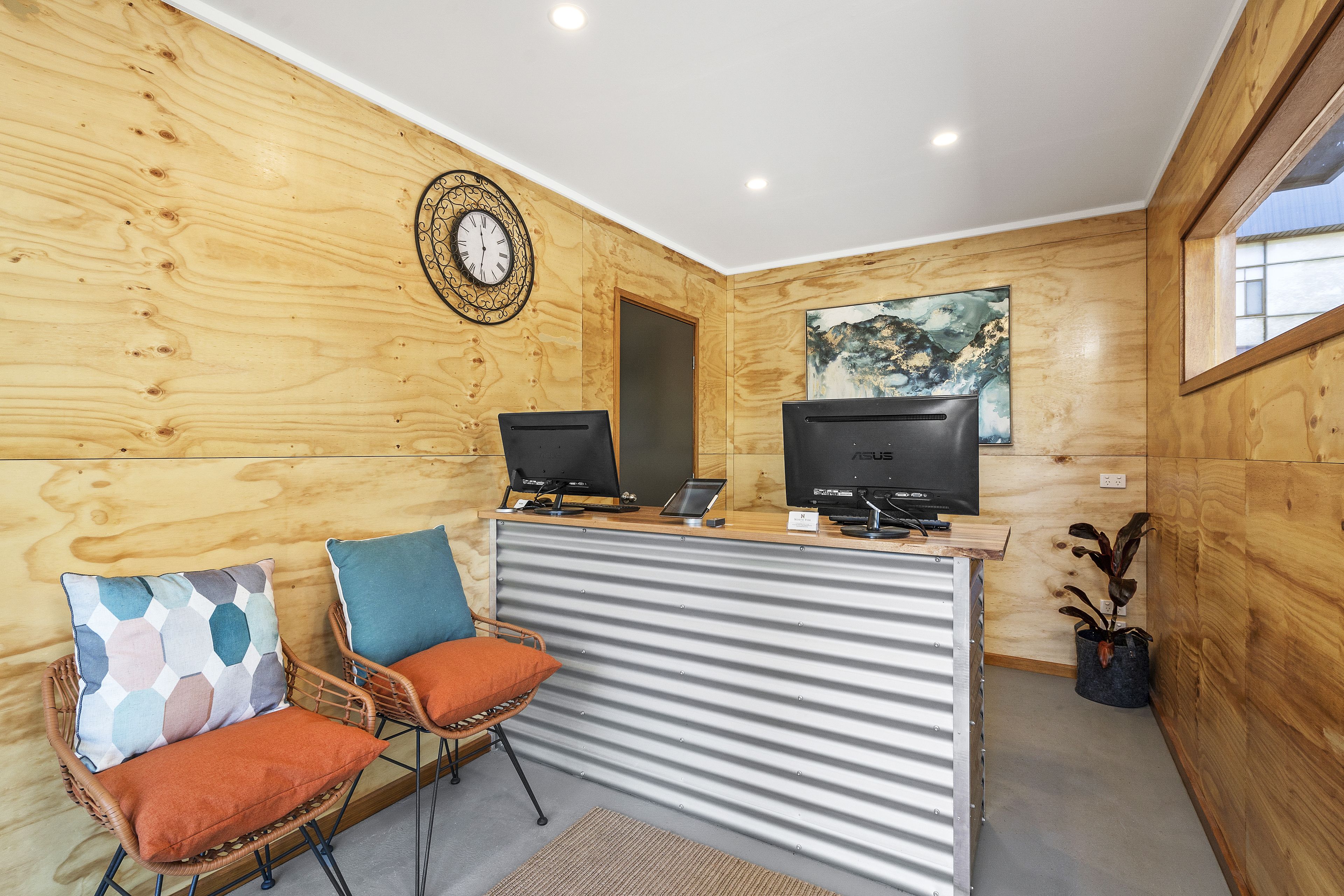 Een receptiegedeelte met houten lambrisering, twee computerschermen, een klok en een schilderij aan de muur.