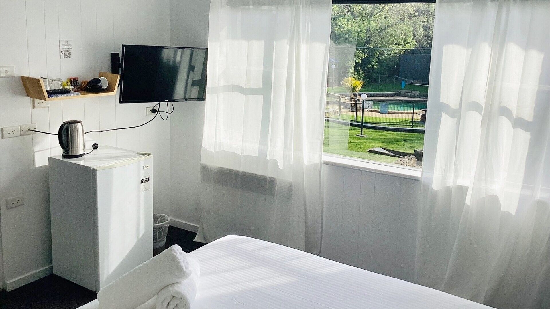 A hotel room with a bed, a television mounted on the wall, a kettle, and a window with sheer curtains.