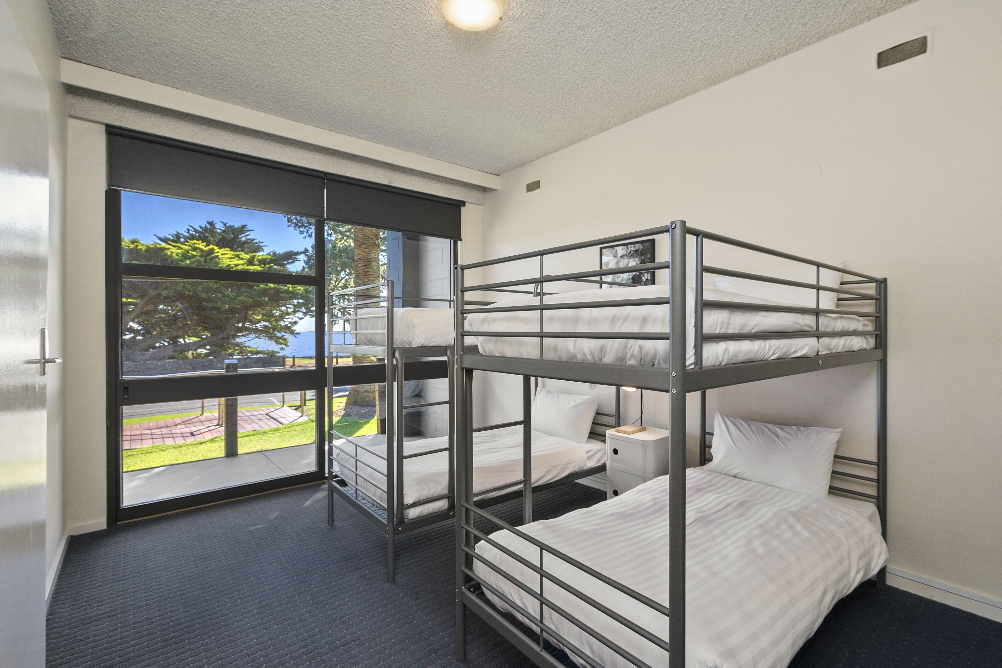 A bunk room with three beds, a window with a view of greenery, and a small bedside table.