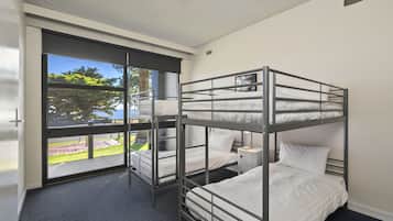 A bunk room with three beds, a window with a view of greenery, and a small bedside table.