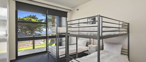 A bunk room with three beds, a window with a view of greenery, and a small bedside table.