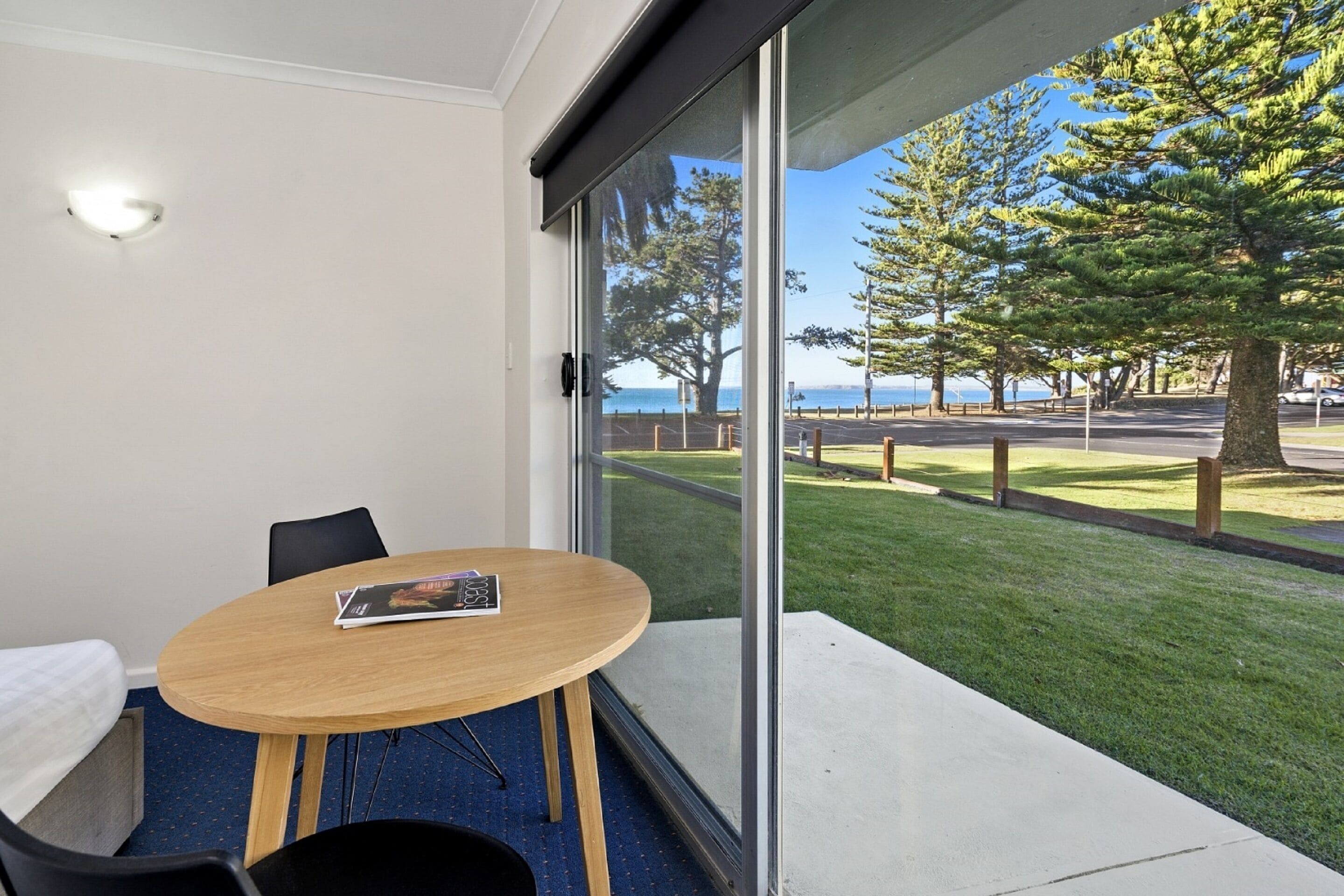 A room with a large window, a round table with chairs, and a view of a grassy area with trees and a body of water.