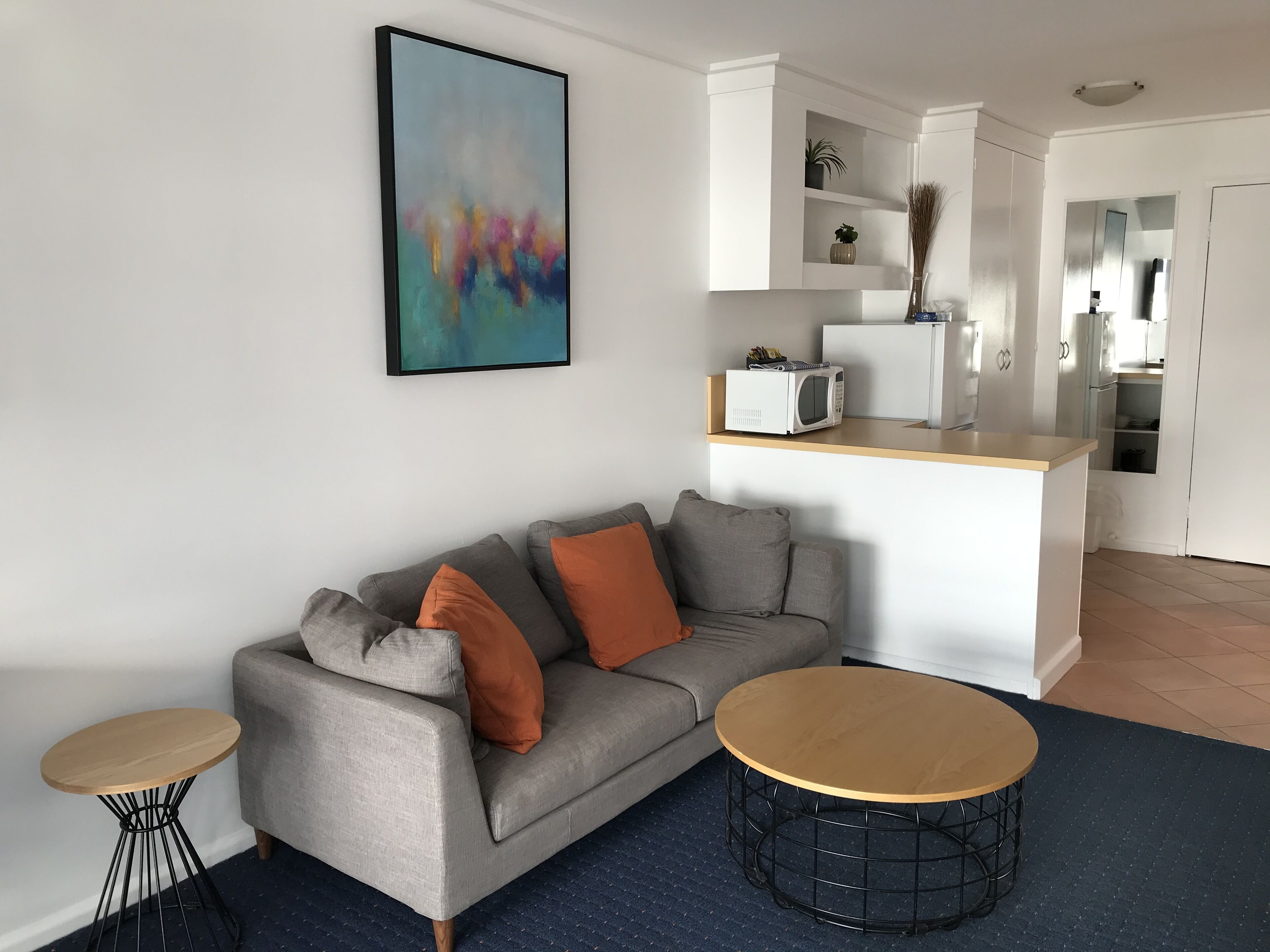 A modern living room with a grey sofa, two round coffee tables, a kitchen area with white cabinets, and a colorful abstract painting on the wall.